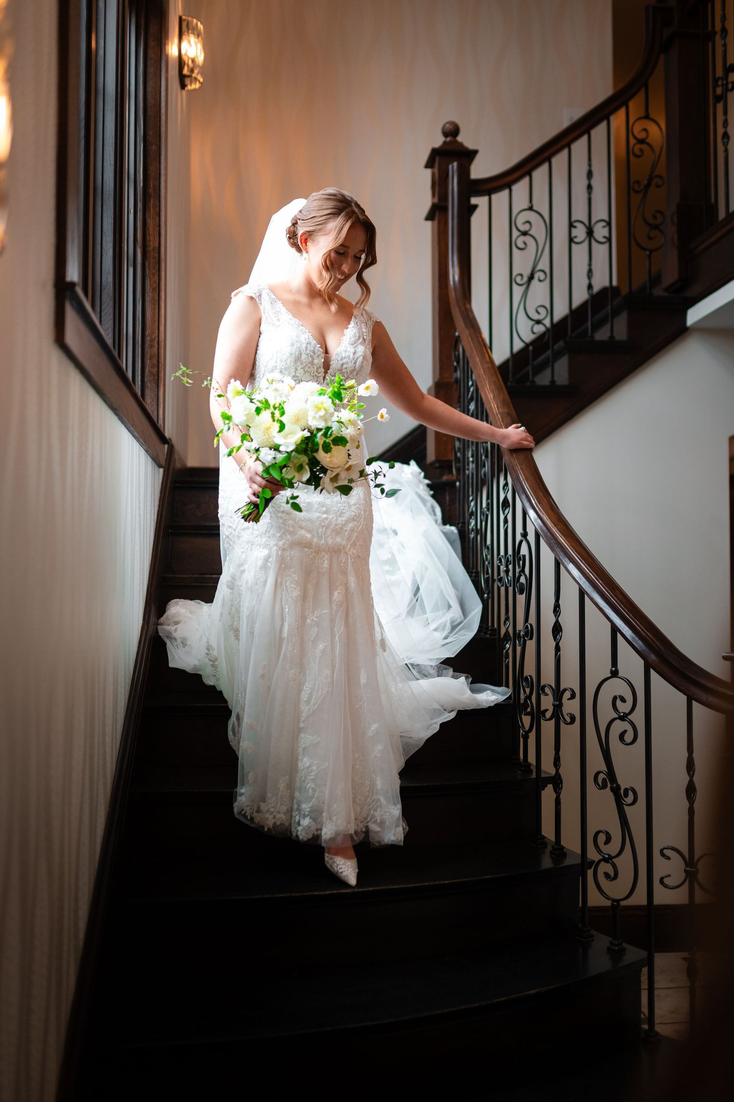 Bride in lace mermaid gown holding white floral bouquet descending elegant curved staircase with iron railing.