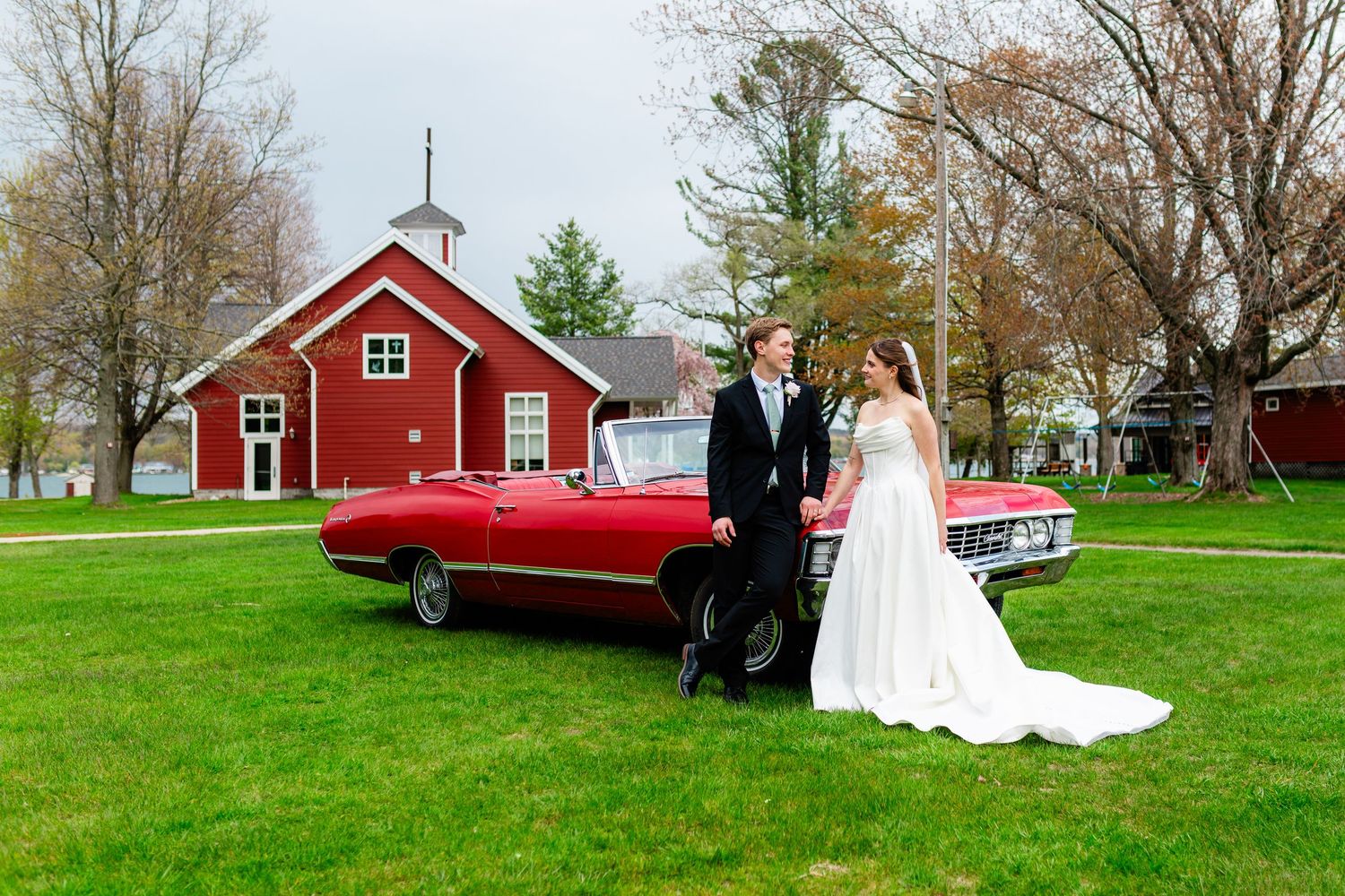 Bride and groom posing beside a red vintage convertible on a green lawn with a charming red barn chapel in the background.