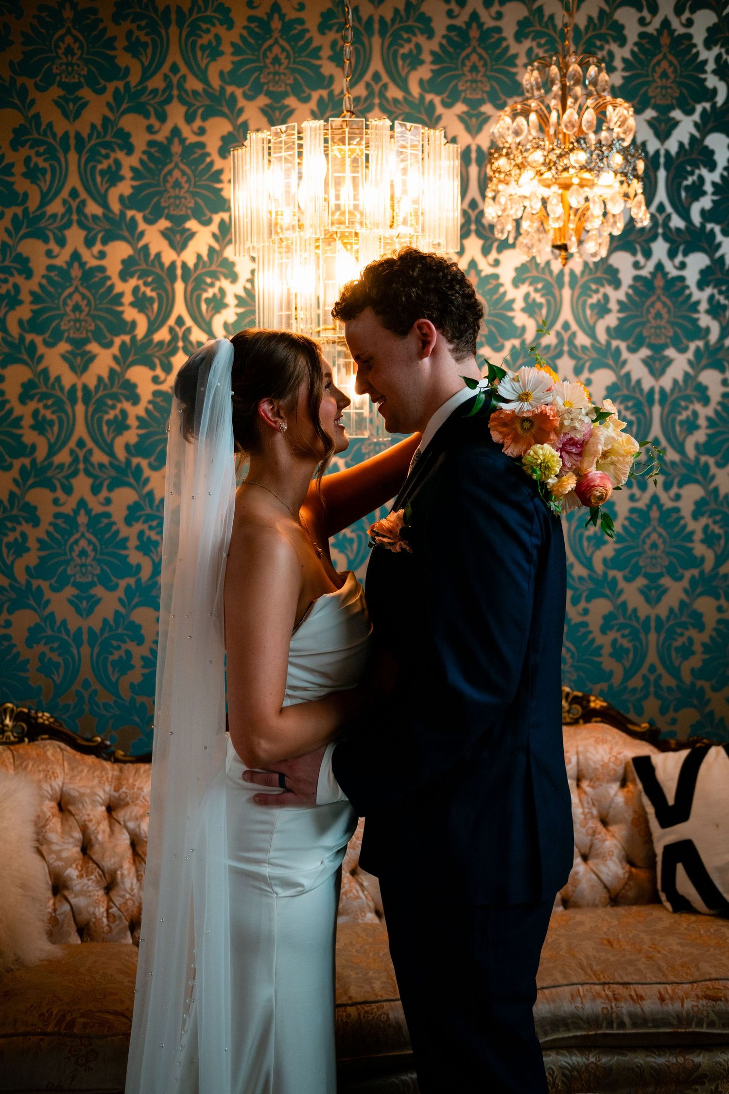 Bride and groom share romantic moment under crystal chandelier, teal damask wallpaper backdrop, wedding bouquet visible.