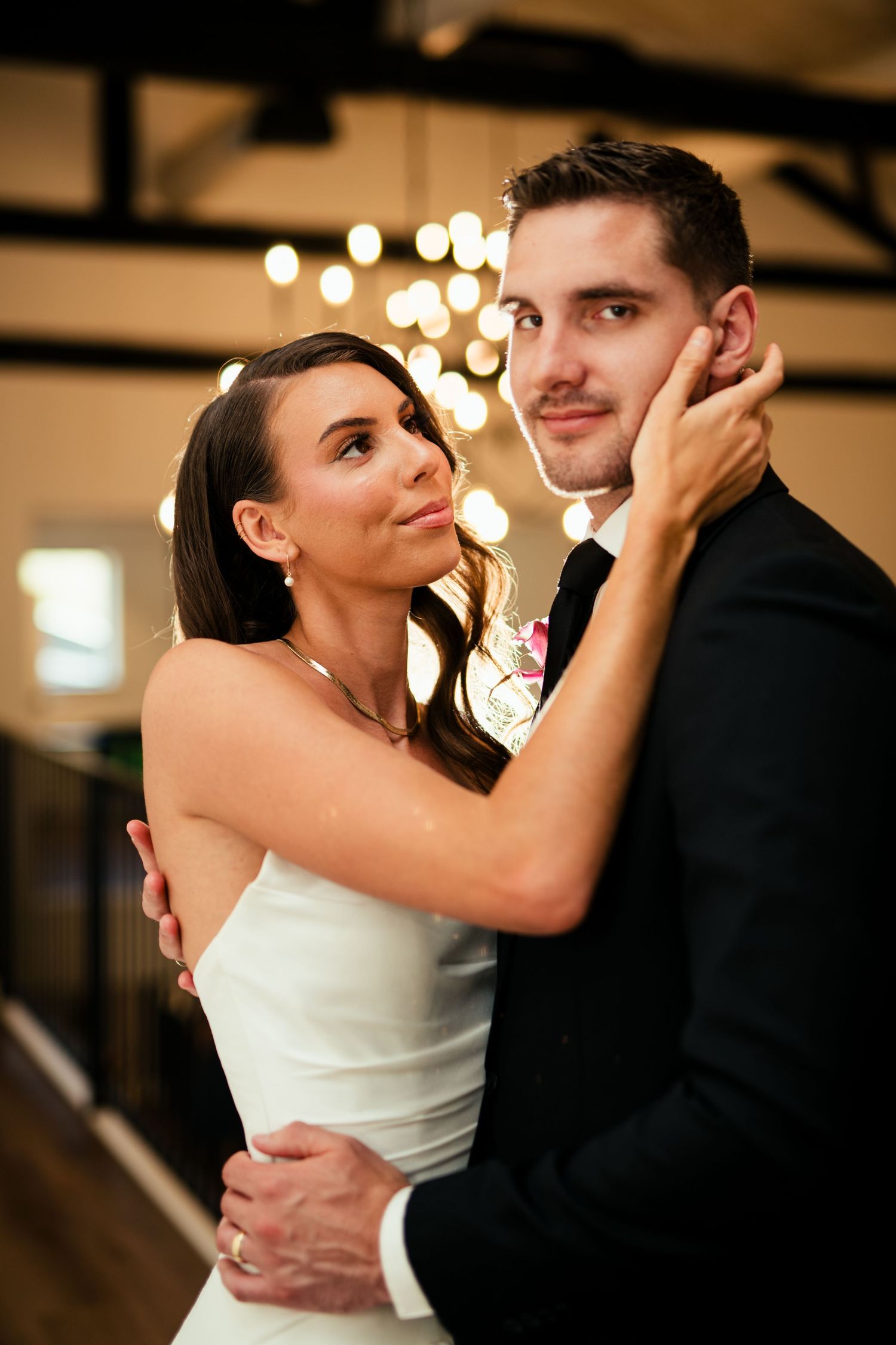 A bride in a white dress and groom in black suit share an intimate moment on their wedding day with soft lights in background.