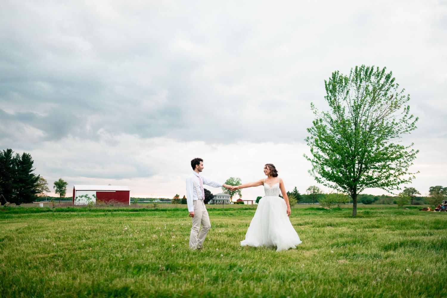 Two figures in wedding attire walk through a green field under a cloudy sky.