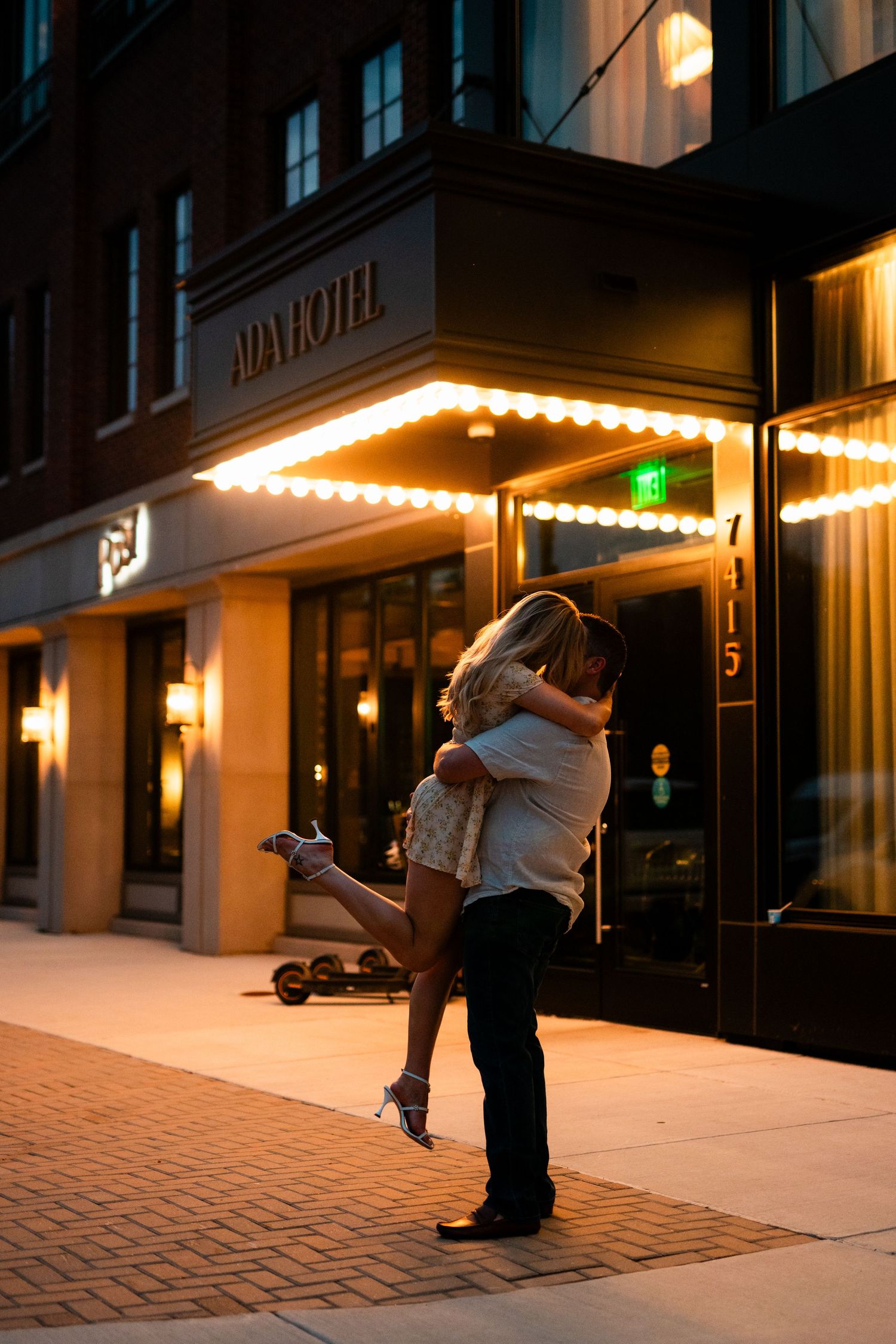 Couple embracing and kissing outside the Ada Hotel at night, lit by warm marquee lights on a city sidewalk.