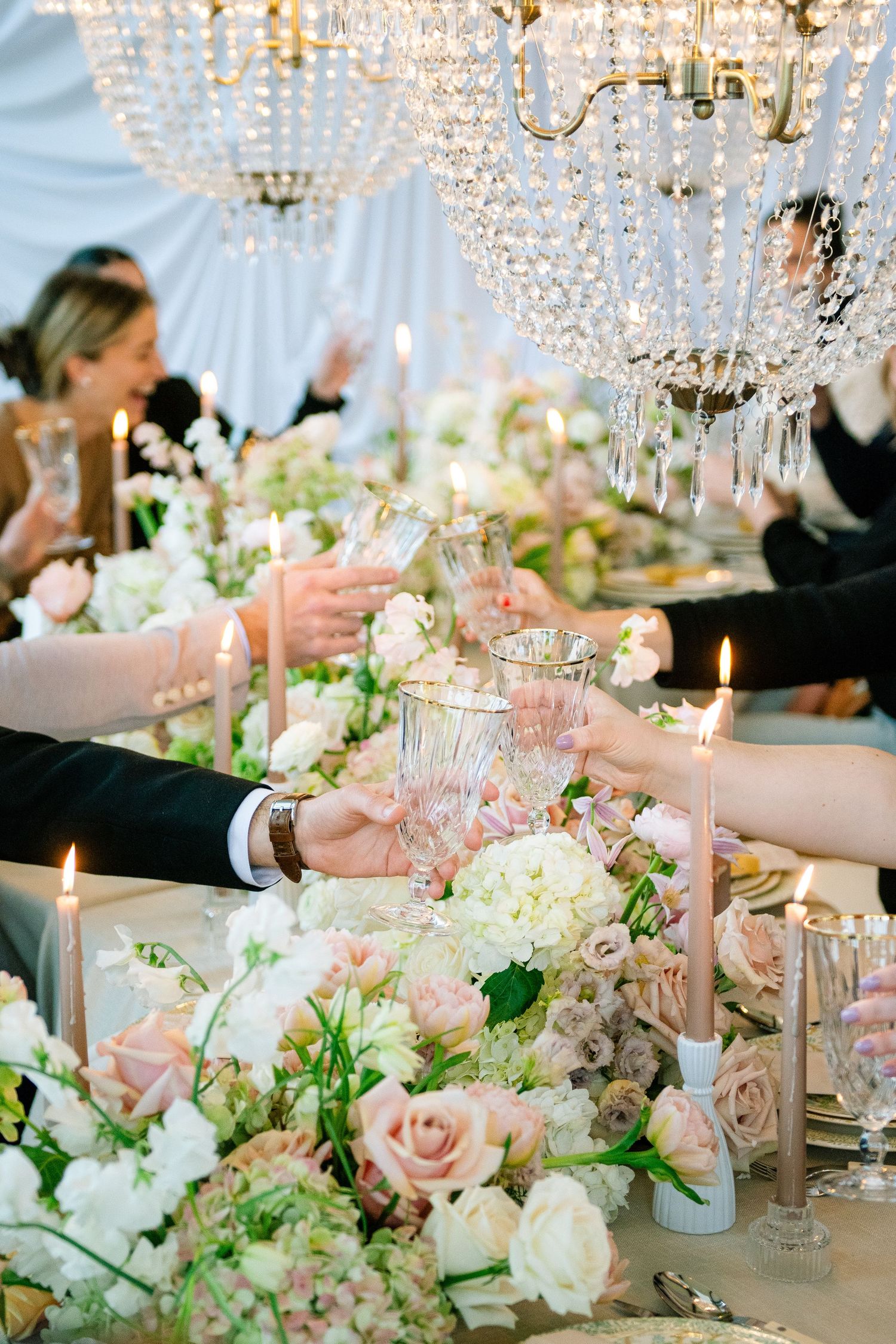 Elegant wedding table setting with crystal chandeliers and blush pink floral centerpieces featuring roses and white flowers.