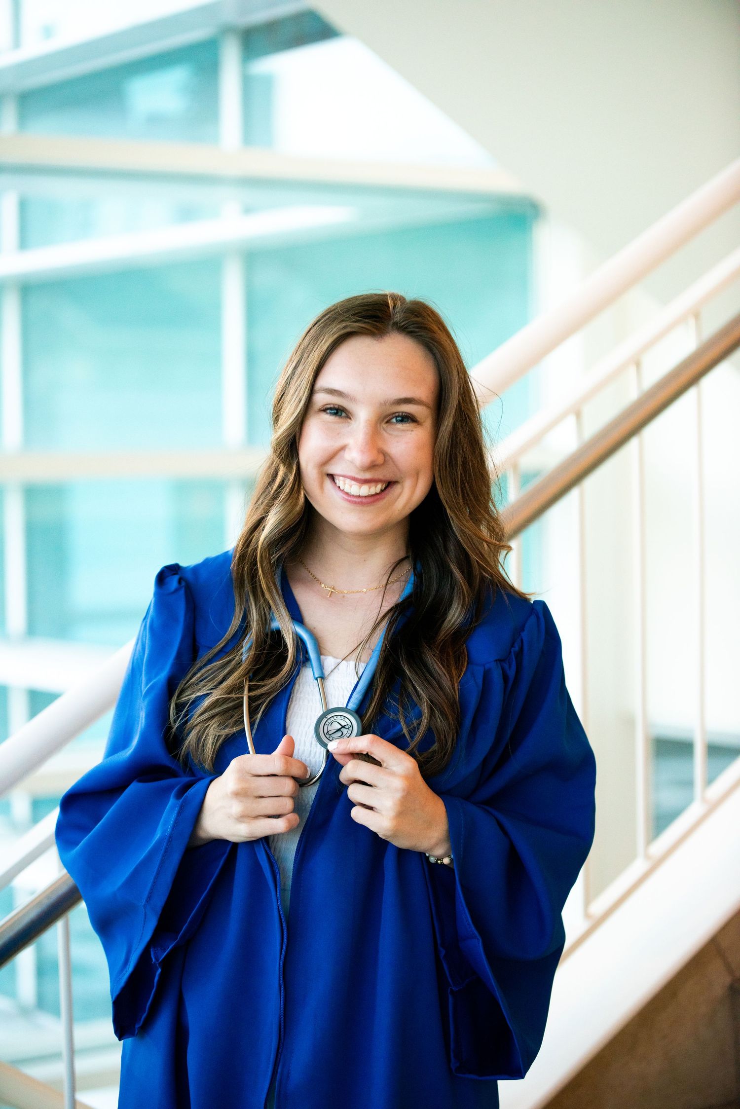 Graduate in bright blue cap and gown smiles while holding stethoscope in modern university building hallway.