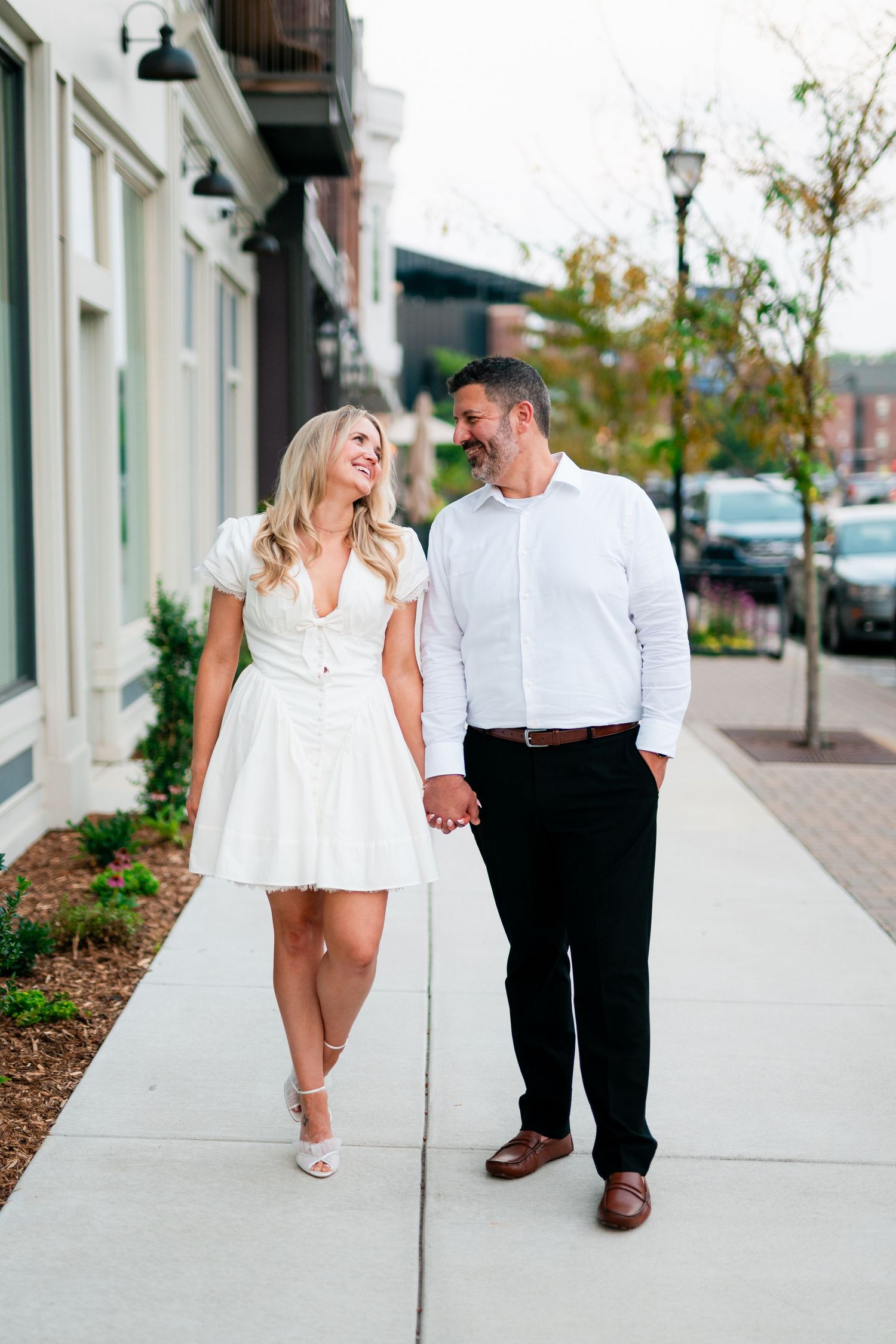 A smiling couple holds hands walking on a sidewalk, woman in white dress, man in white shirt and black pants.