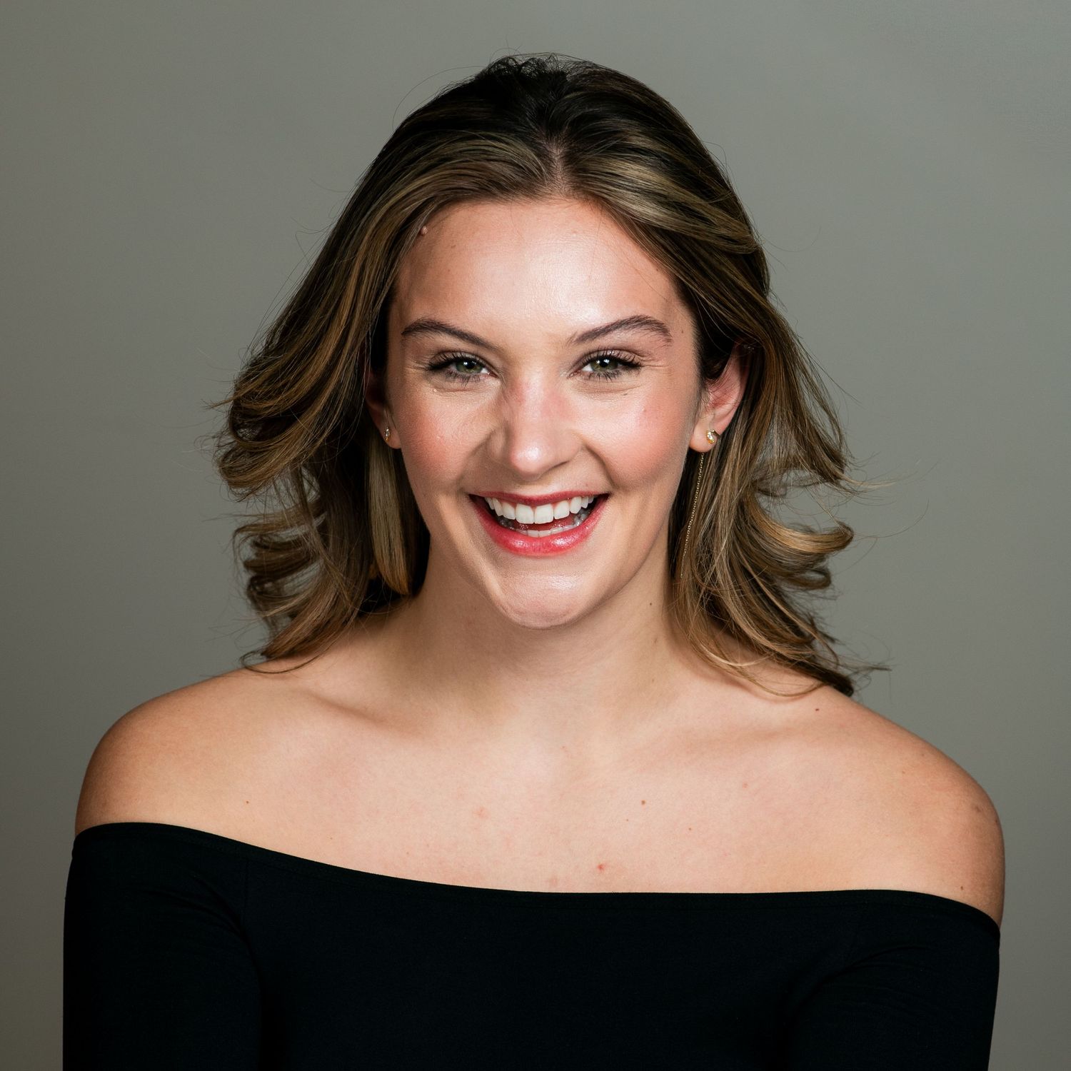 A smiling woman with wavy brown hair wearing an off-shoulder black top poses against a gray backdrop.