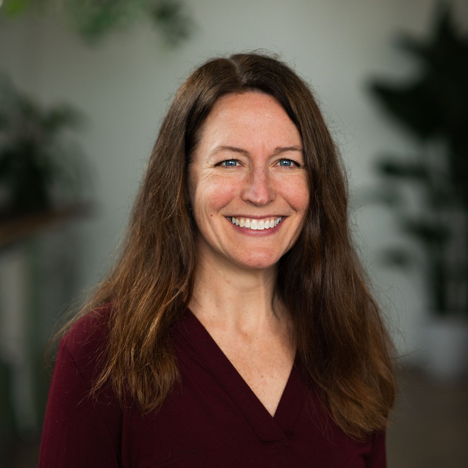 Professional headshot of someone with long brown hair and a warm smile wearing a burgundy v-neck top.