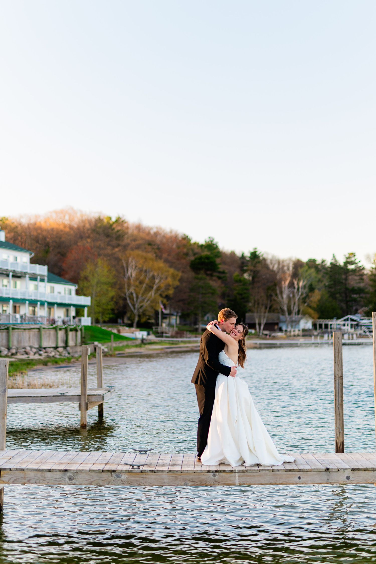 Bride and groom embrace on a lakeside dock at golden hour, with trees and resort buildings in the background.