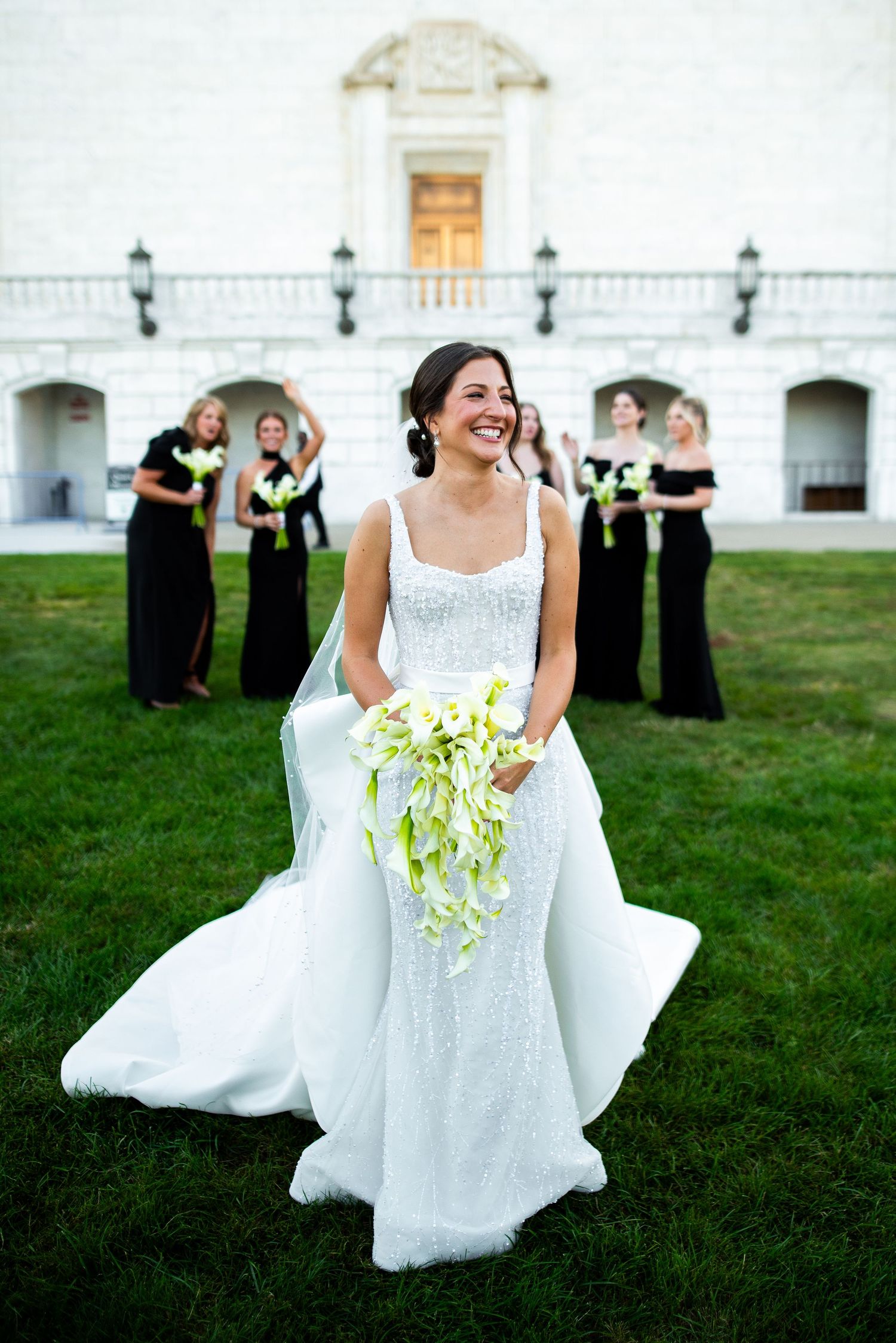 A bride in a white gown twirls on grass while bridesmaids in black dresses stand in the background.