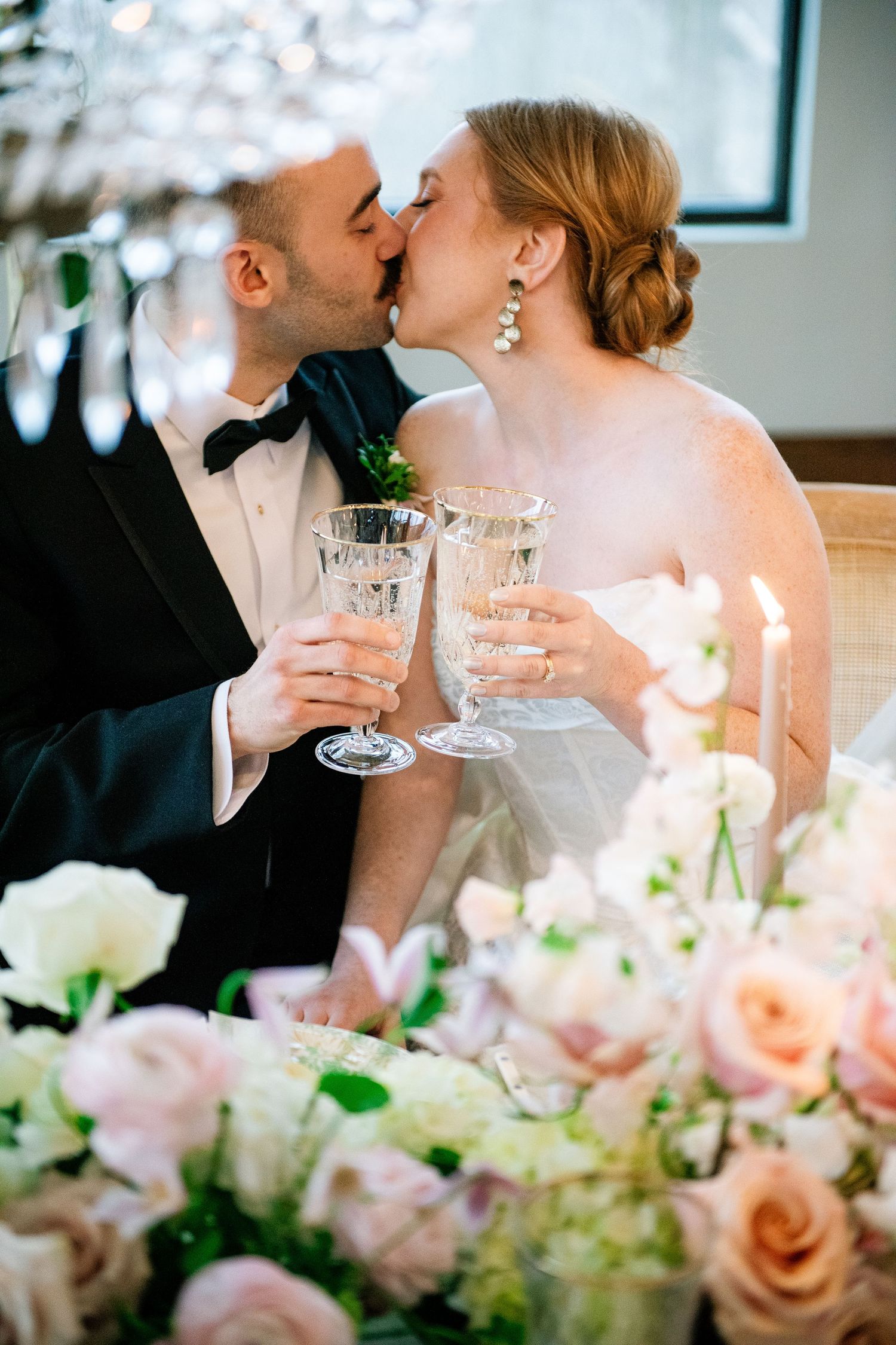 Newlyweds share romantic champagne toast at reception table decorated with pink roses and glowing candles.