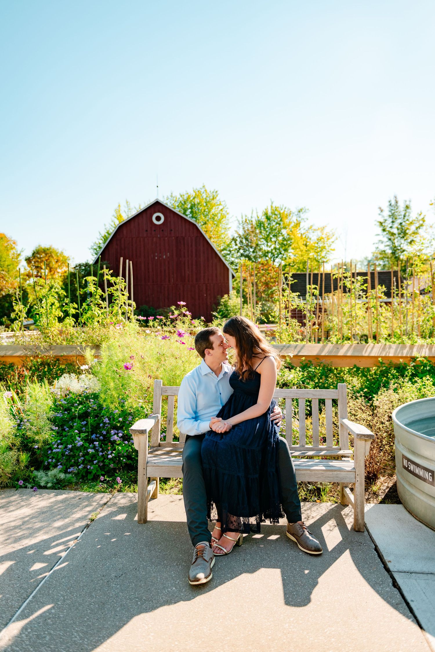 A couple sits together on a wooden bench in a garden setting with a red barn and autumn foliage in the background.