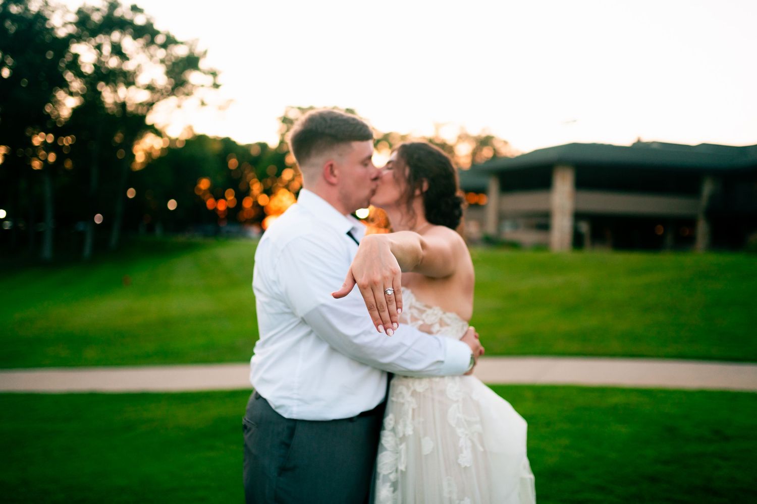 A romantic couple embraces and kisses on a green lawn during a wedding celebration.