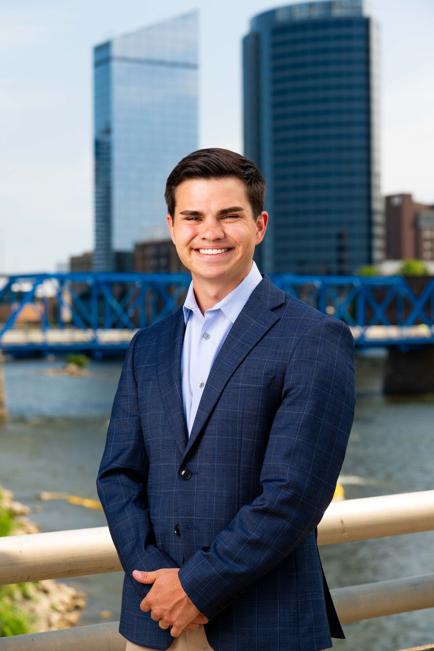Young professional man in navy blazer smiling outdoors by a river with a blue bridge and city skyline.
