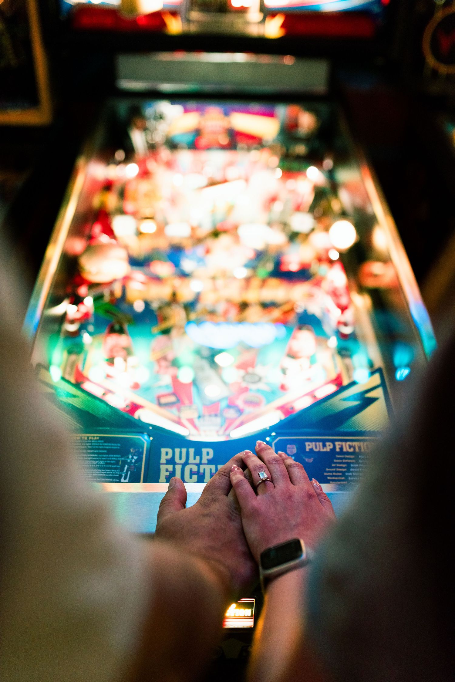 Hands on a Pulp Fiction pinball machine with colorful glowing lights in an arcade setting.