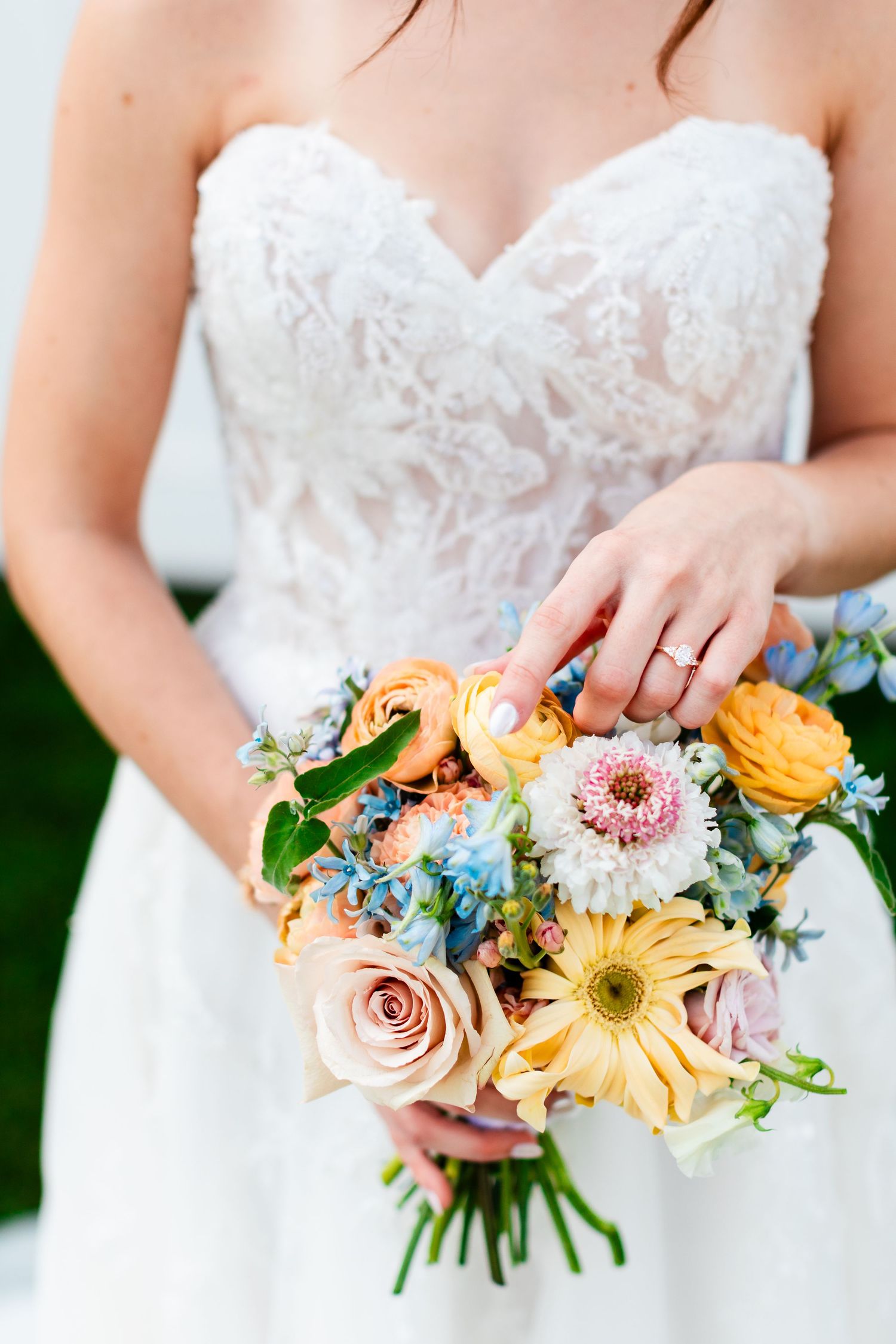 Bride in white lace gown holding colorful wedding bouquet with yellow, pink, and blue flowers.