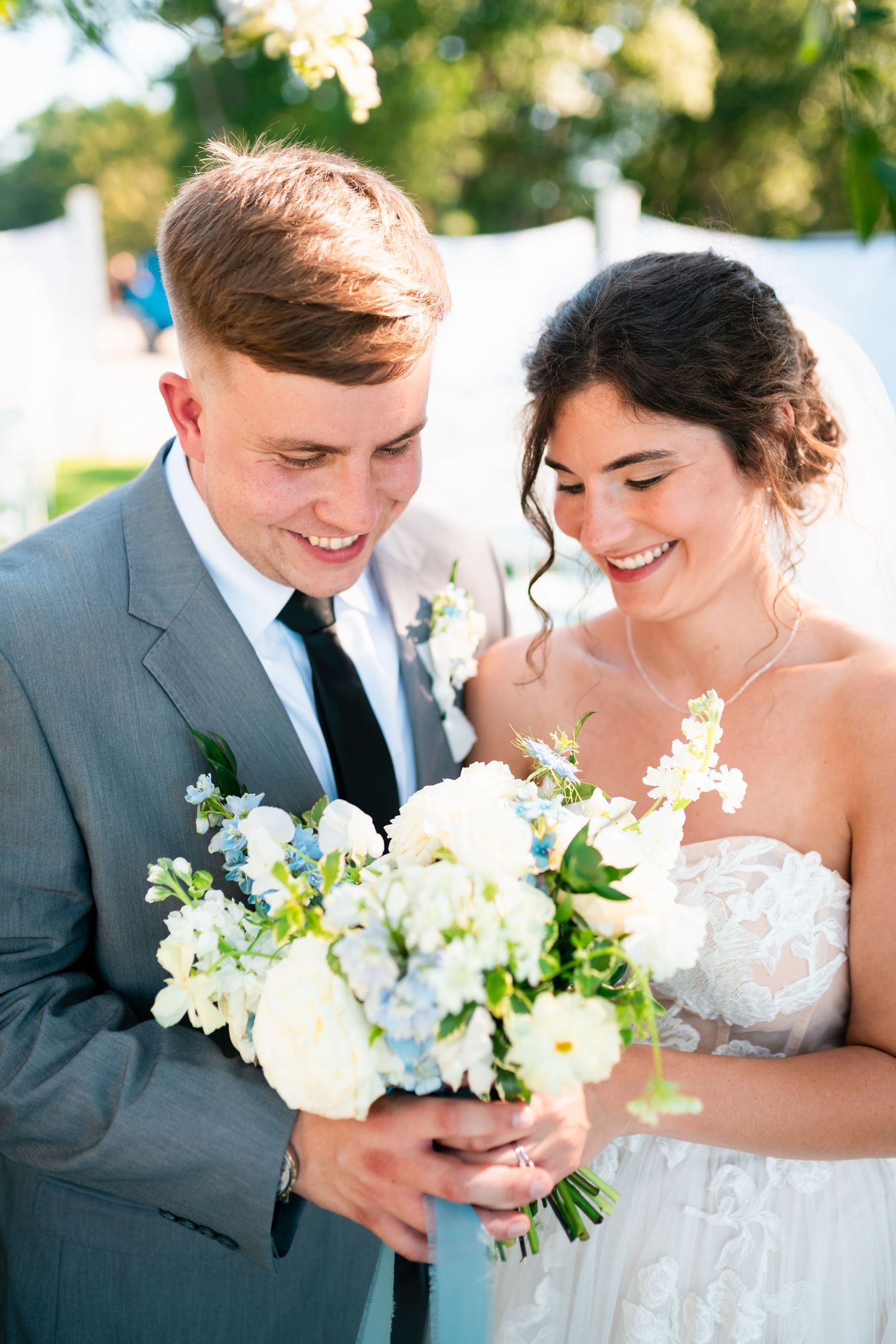 Newlywed couple share intimate moments while holding white floral bouquet in outdoor wedding setting.