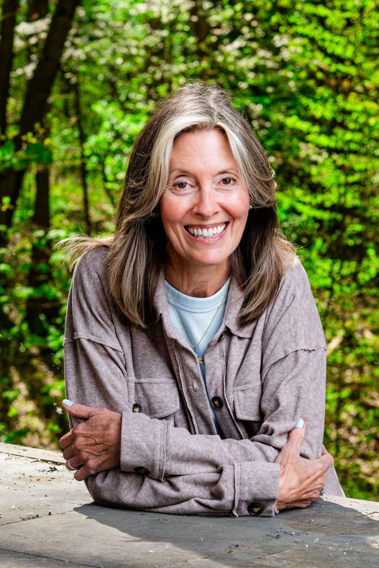 Smiling middle-aged woman with long gray hair leaning on a stone surface outdoors surrounded by green trees.