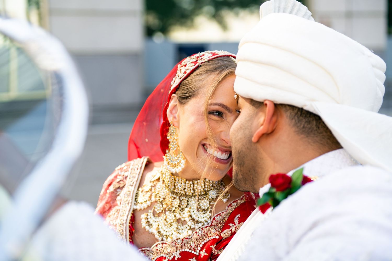 Smiling bride in red lehenga and gold jewelry with groom in white turban at a traditional Indian wedding.