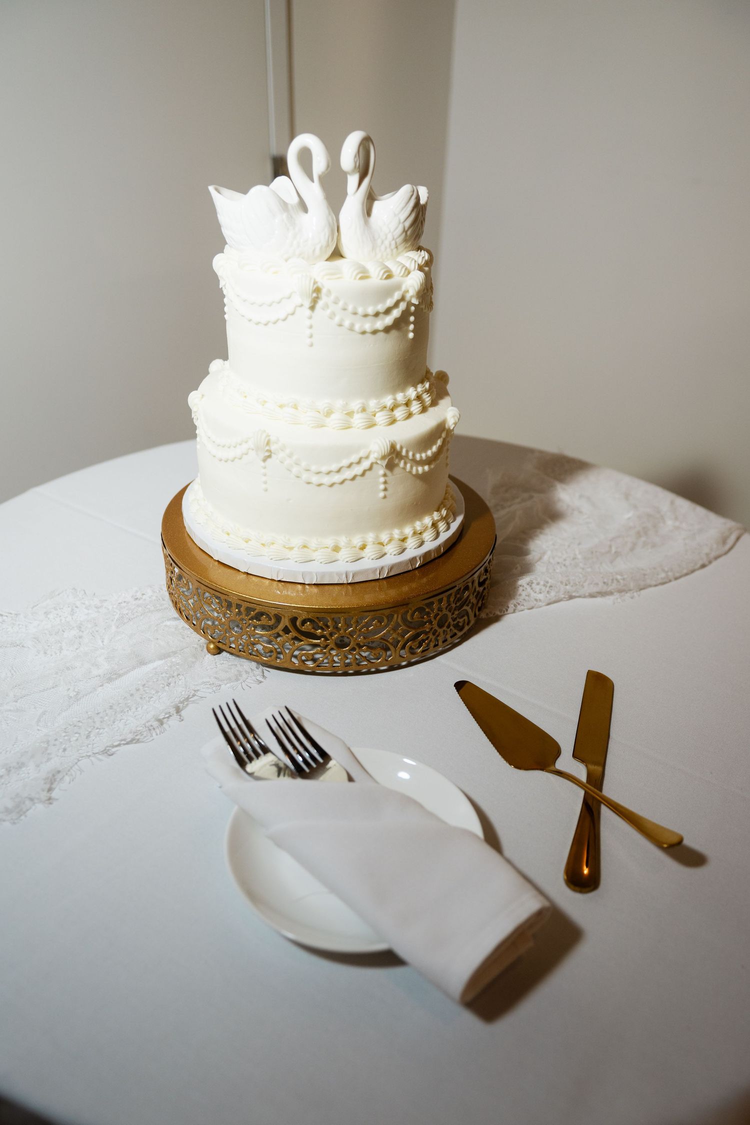 Elegant two-tier white wedding cake with swan toppers on gold stand, beside gold cake server set.