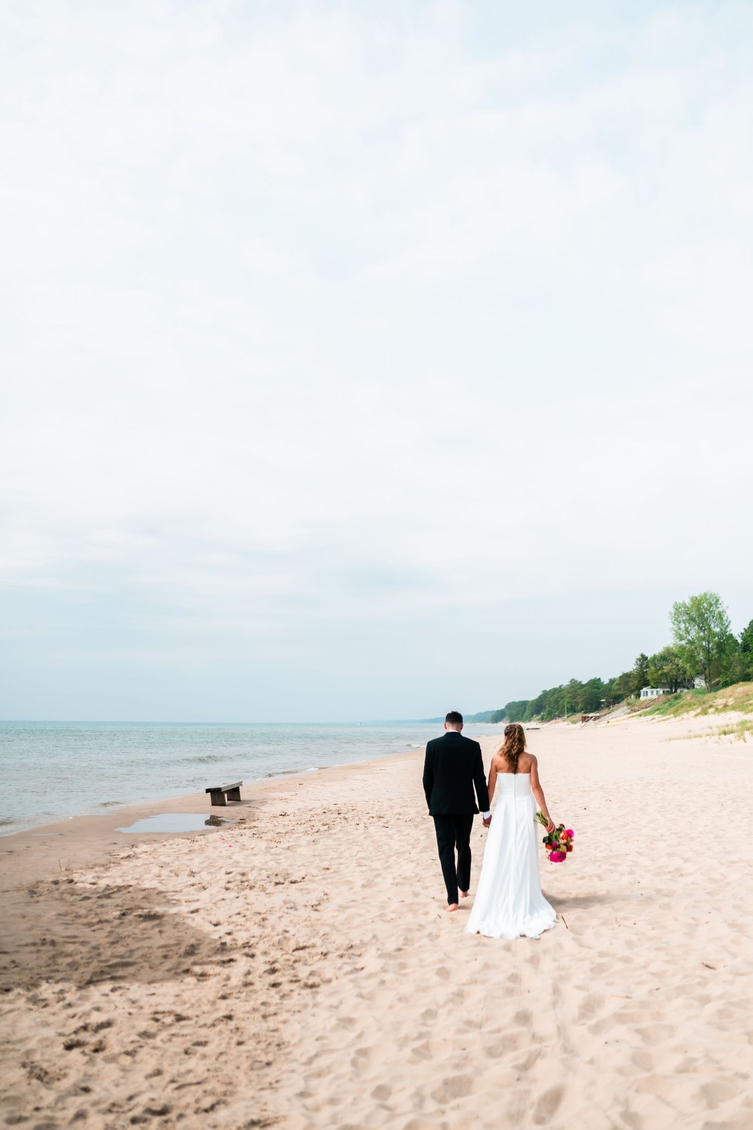 A couple in wedding attire walks along a serene beach shoreline with ocean views and sandy landscape.