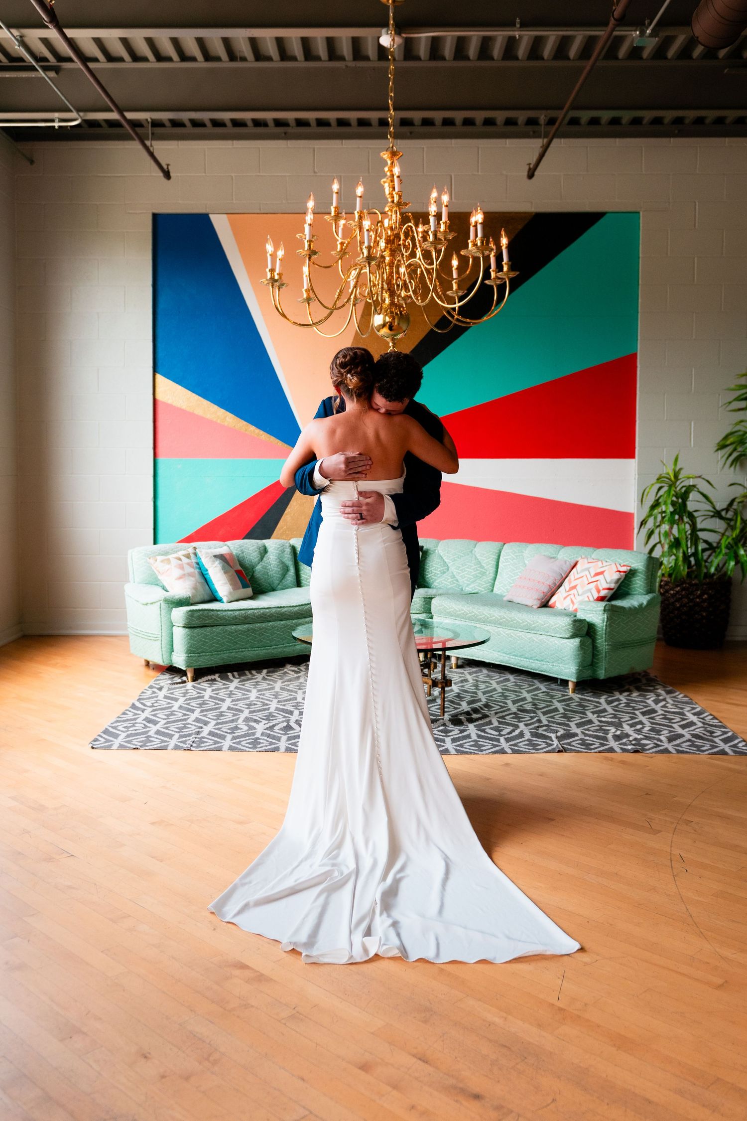 Elegant wedding scene with couple embracing in front of colorful geometric wall art and mint green sofa under chandelier.