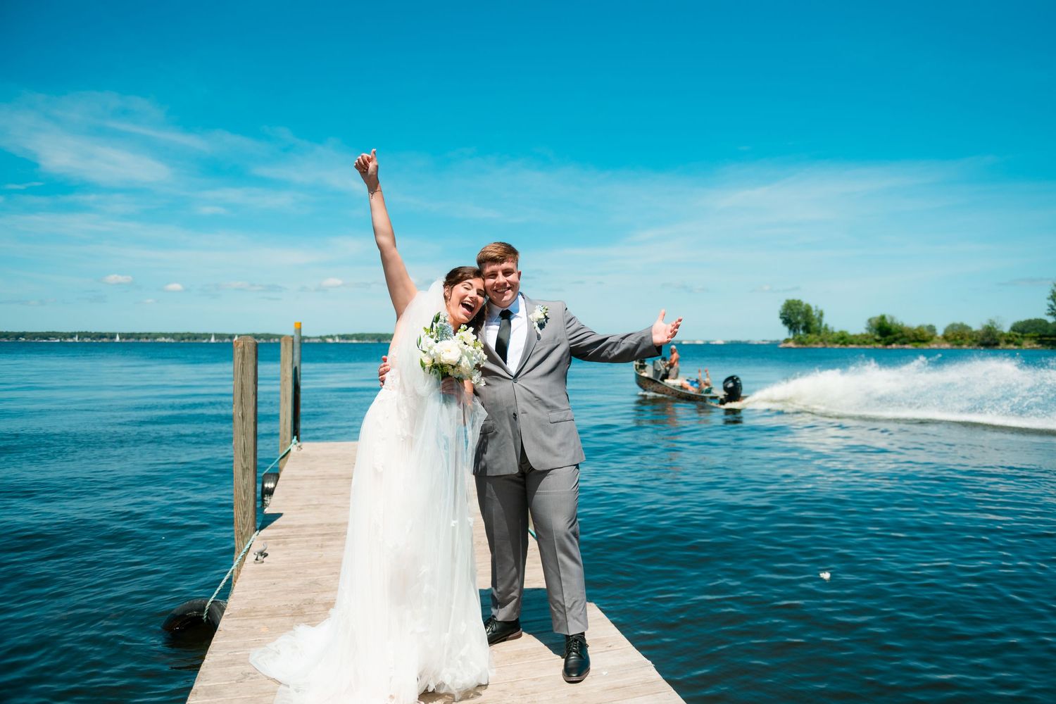 Couple in wedding attire embraces on a wooden dock overlooking a serene blue lake on a sunny summer day.