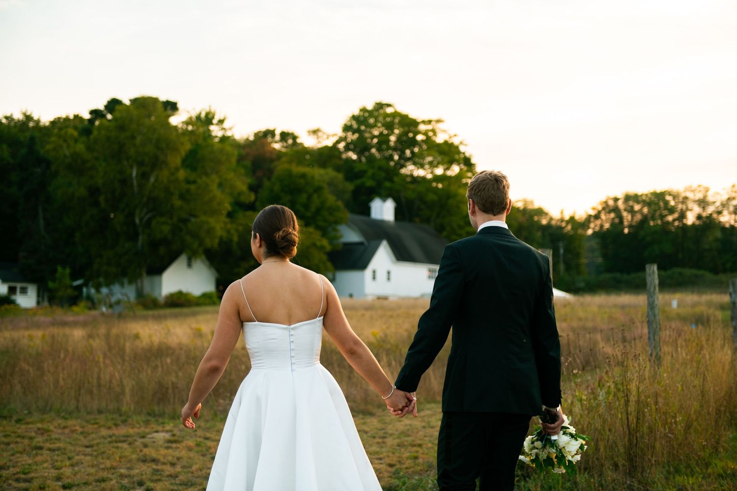 Bride and groom holding hands walking toward a white farmhouse at sunset in a golden field.