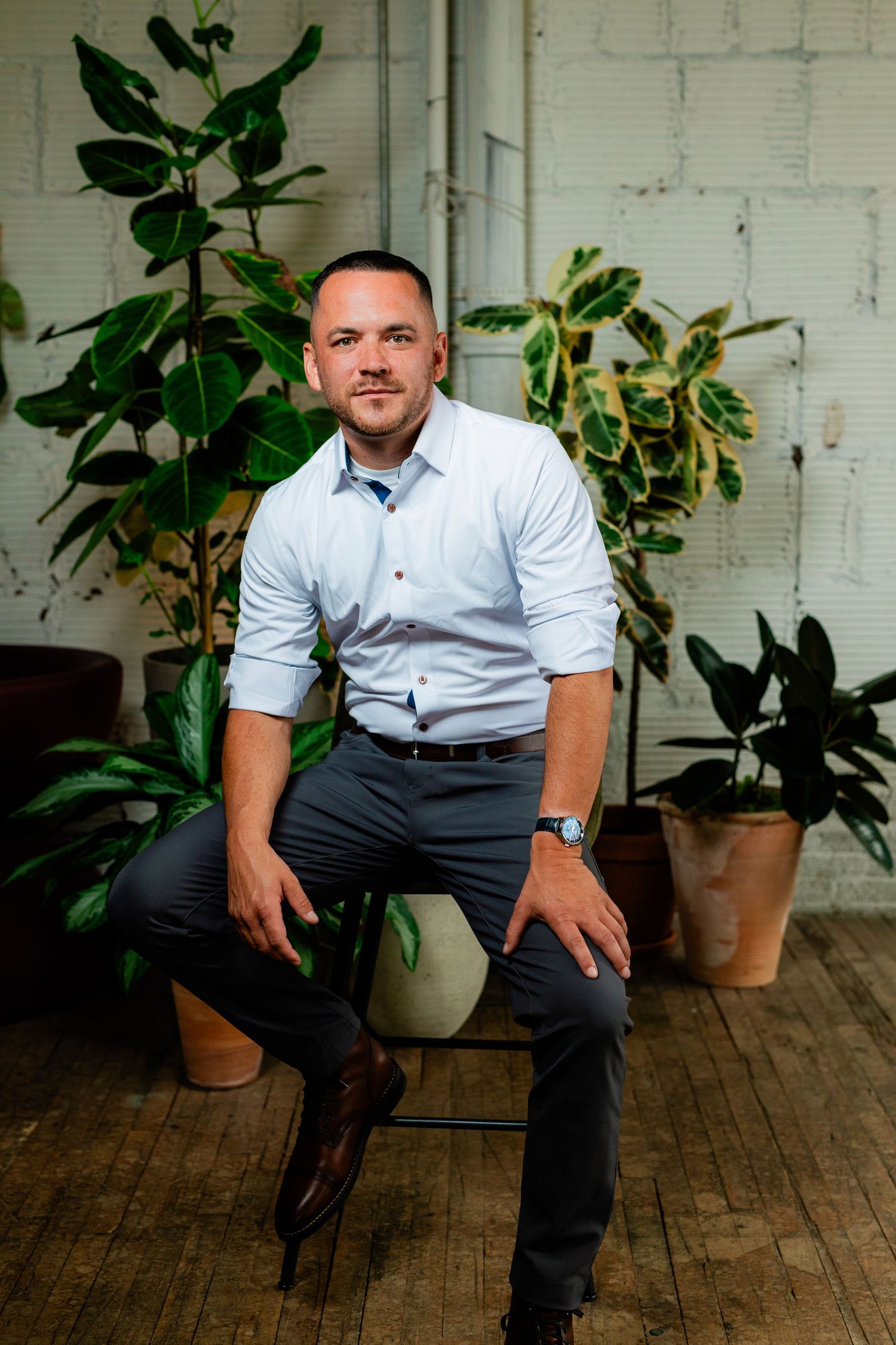 Man in light blue shirt and dark pants sits on stool surrounded by lush green potted plants in rustic studio.