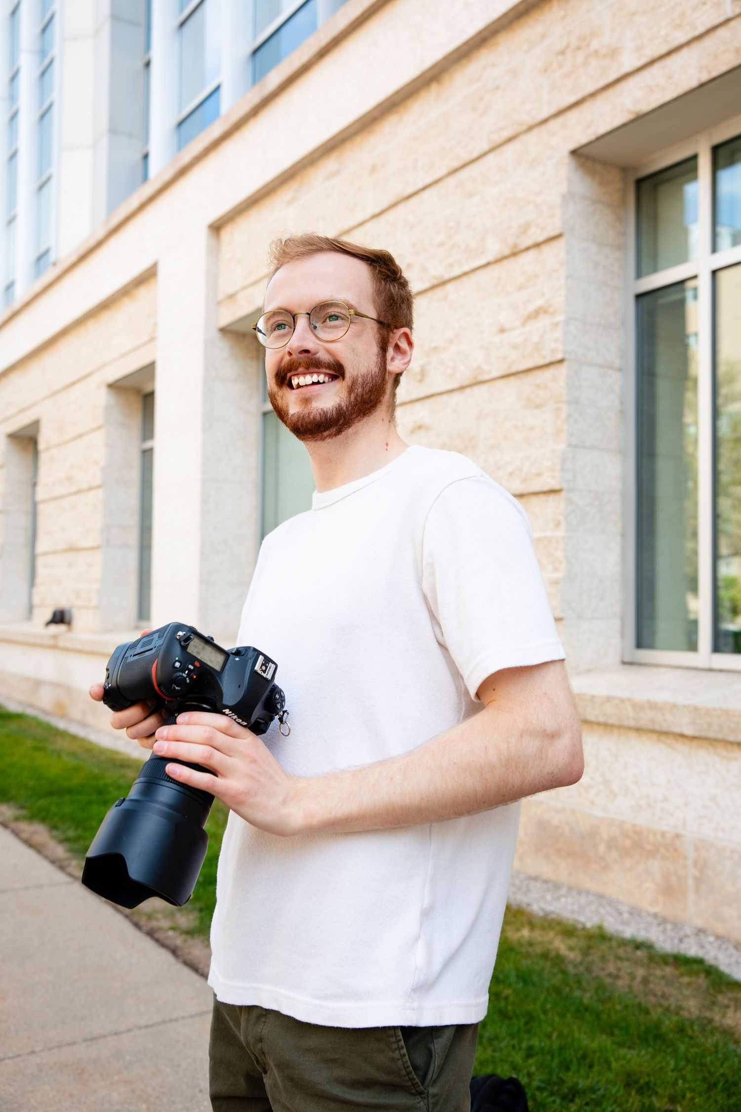 A photographer wearing glasses and a white t-shirt holds a professional DSLR camera with a telephoto lens outdoors.