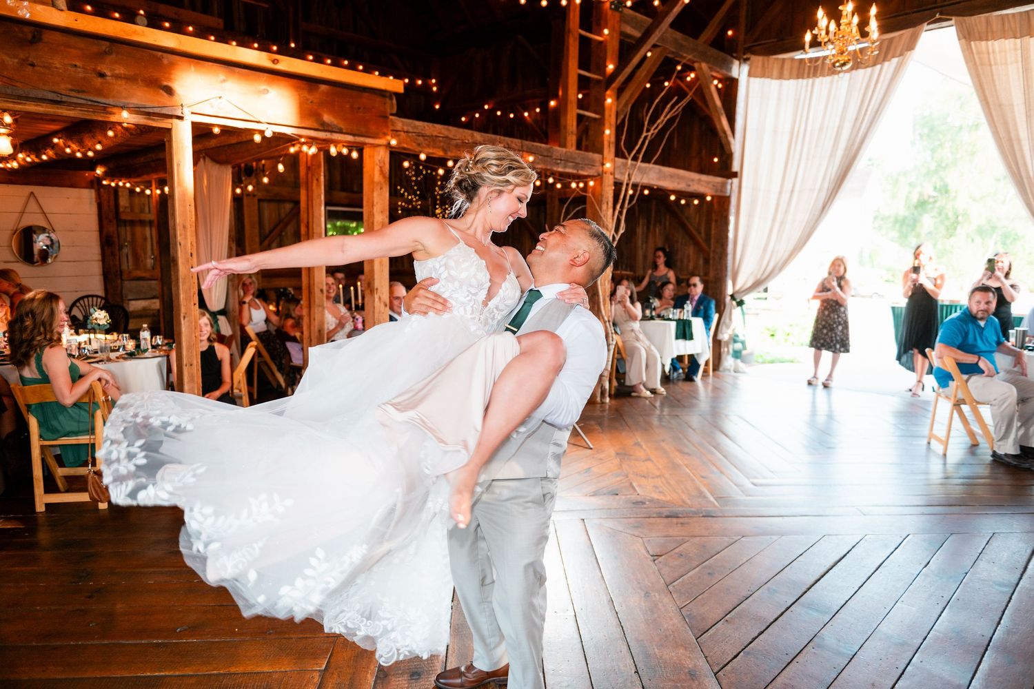 Couple dancing at a rustic barn wedding reception with string lights and white drapes creating a romantic atmosphere.