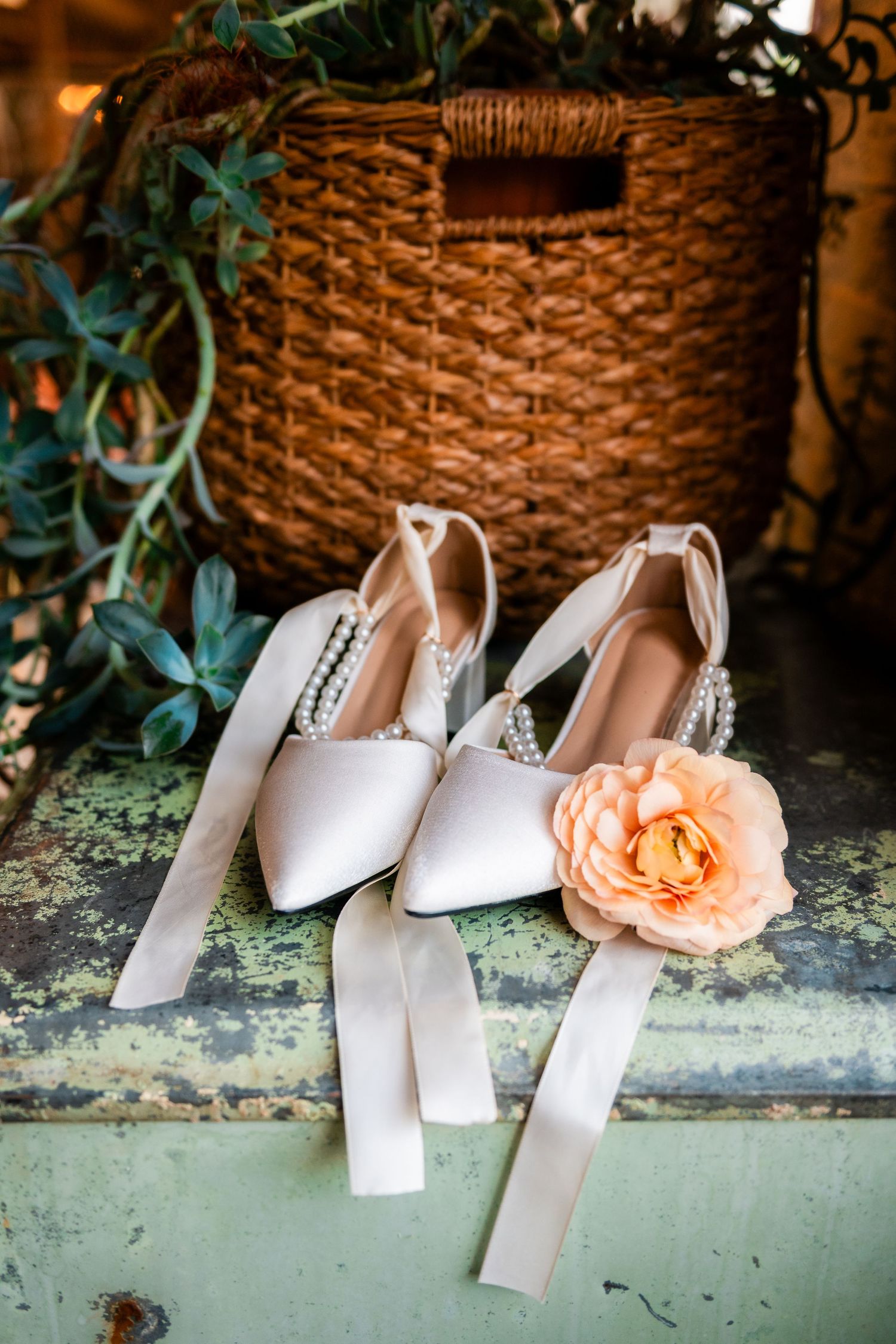 White bridal heels with crystal embellishments and a peach rose accent displayed against a rustic green background.
