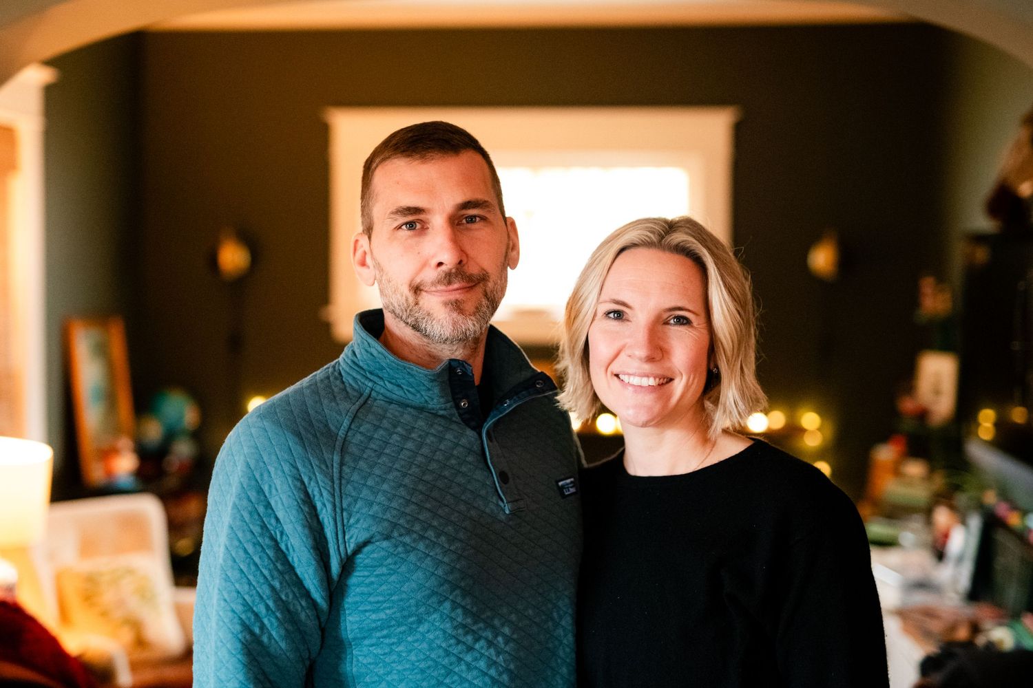 Couple sits together in a warmly lit room wearing casual attire during at-home family lifestyle session in west michigan