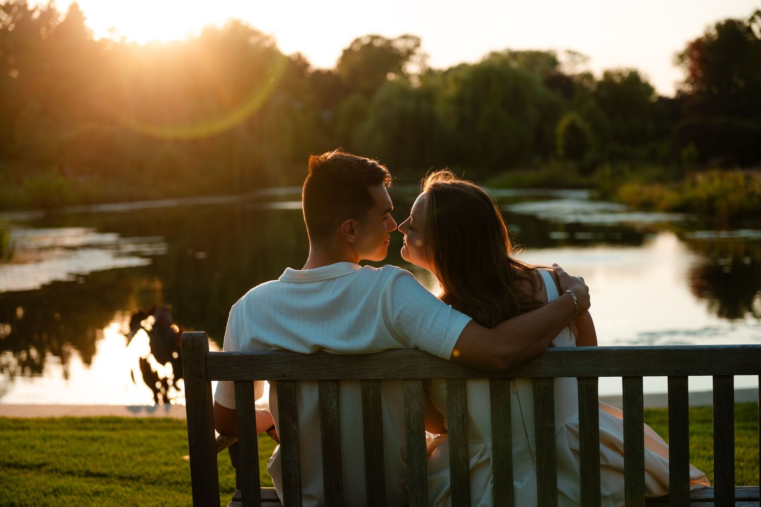 Couple sitting on a wooden bench sharing a romantic moment by a serene pond at golden sunset.