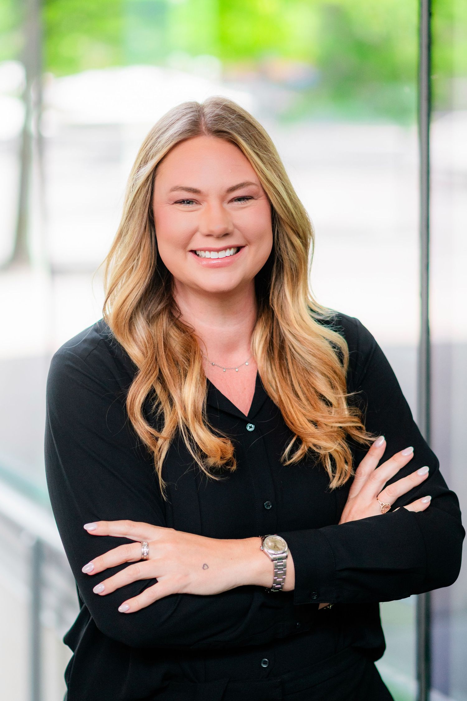Professional headshot of a smiling blonde woman in a black blouse with arms crossed in a modern office setting.