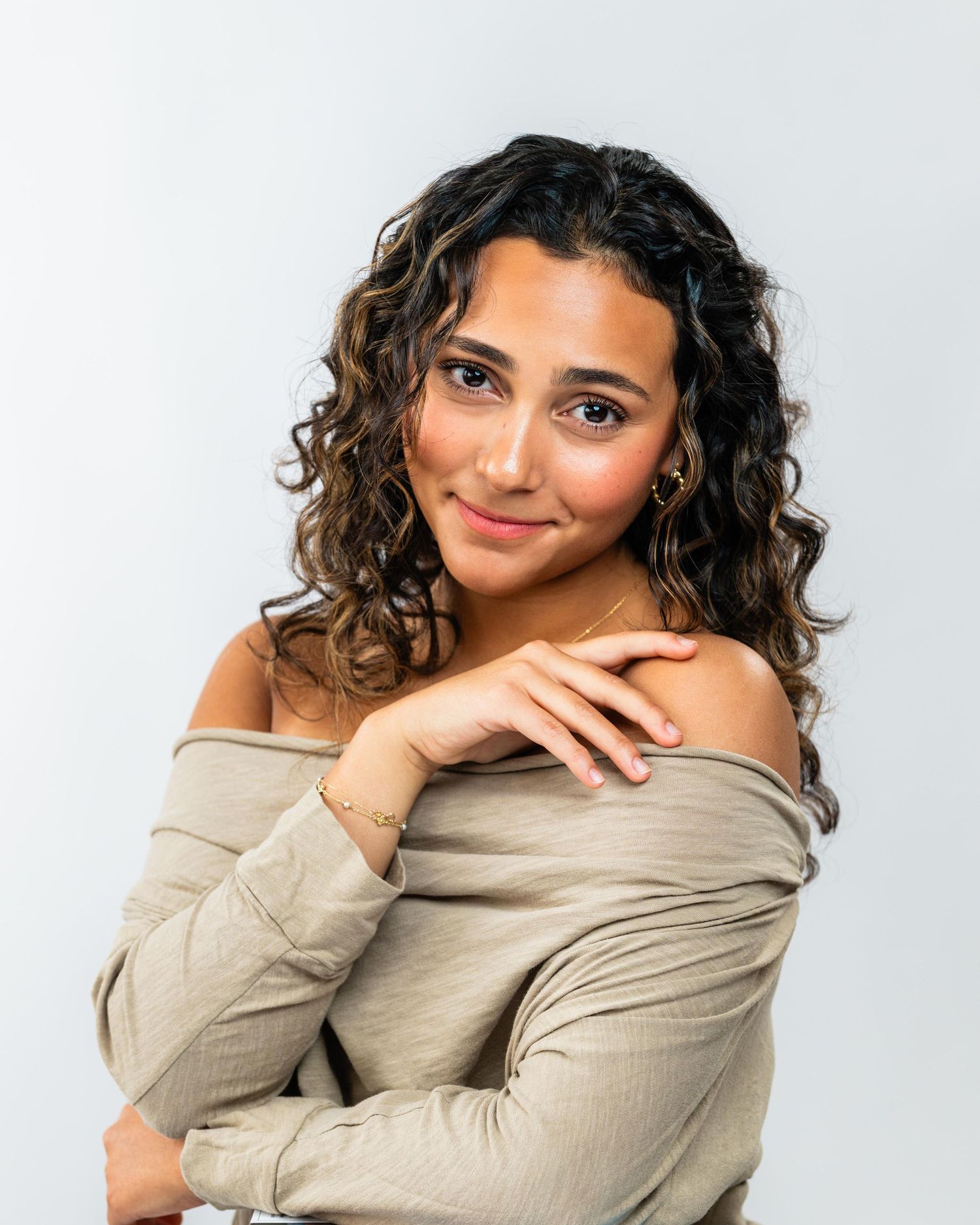 Young woman with curly hair wearing an off-shoulder beige top, posing confidently against a light gray background.