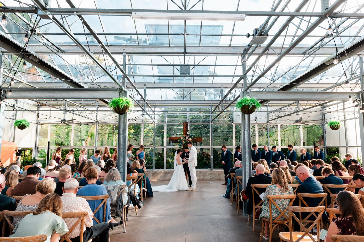 A modern greenhouse wedding ceremony with glass ceiling and wooden chairs filled with guests watching the ceremony.