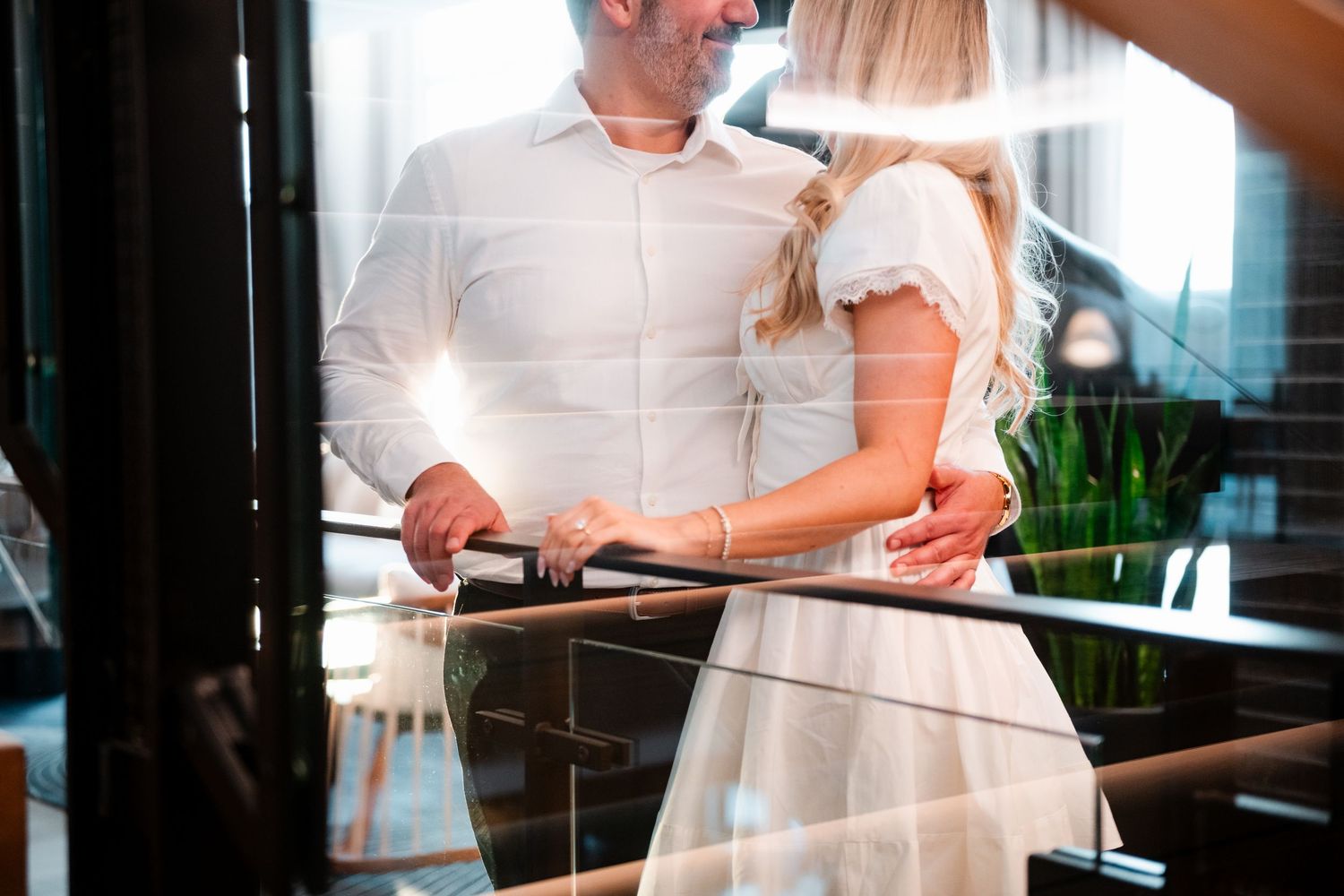 Romantic couple in white outfits embracing near glass railing in elegant indoor setting.