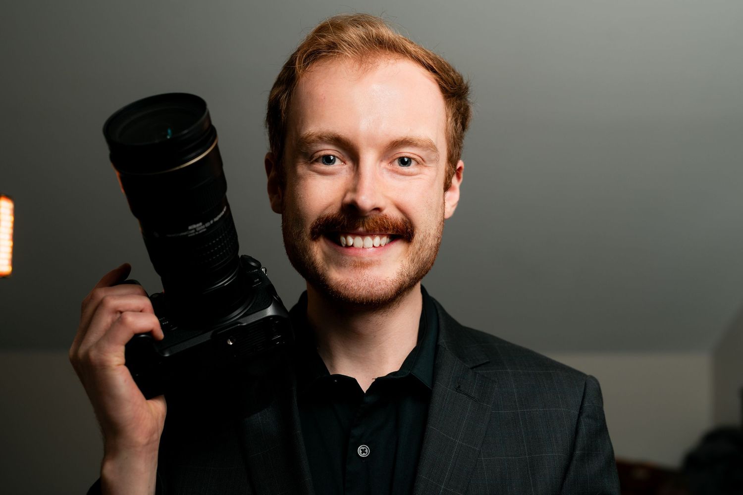 A person wearing a black shirt smiles while holding a camera against a light background.