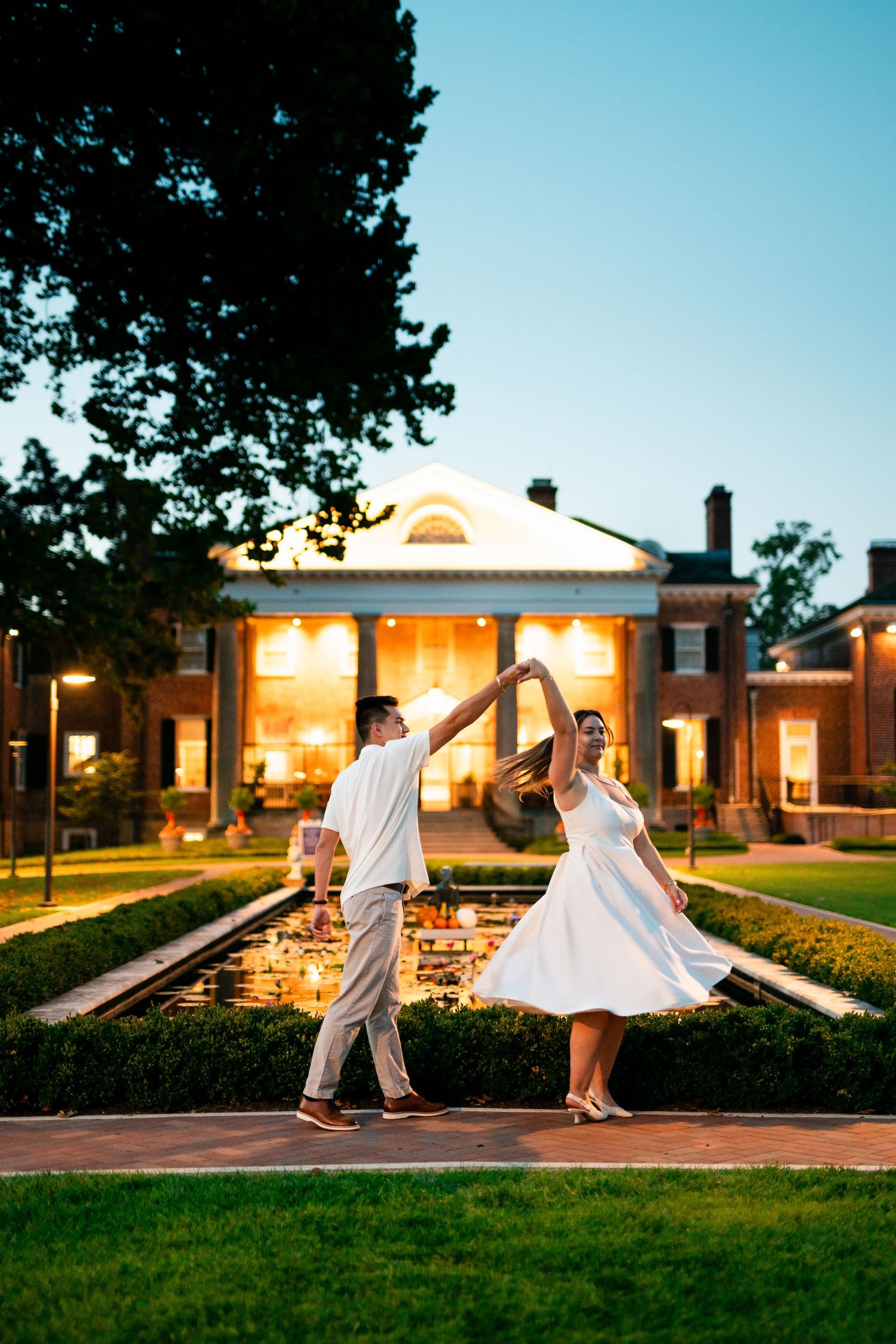 Couple dancing in white outfits by a reflecting pool at dusk, with an elegant lit mansion in the background.