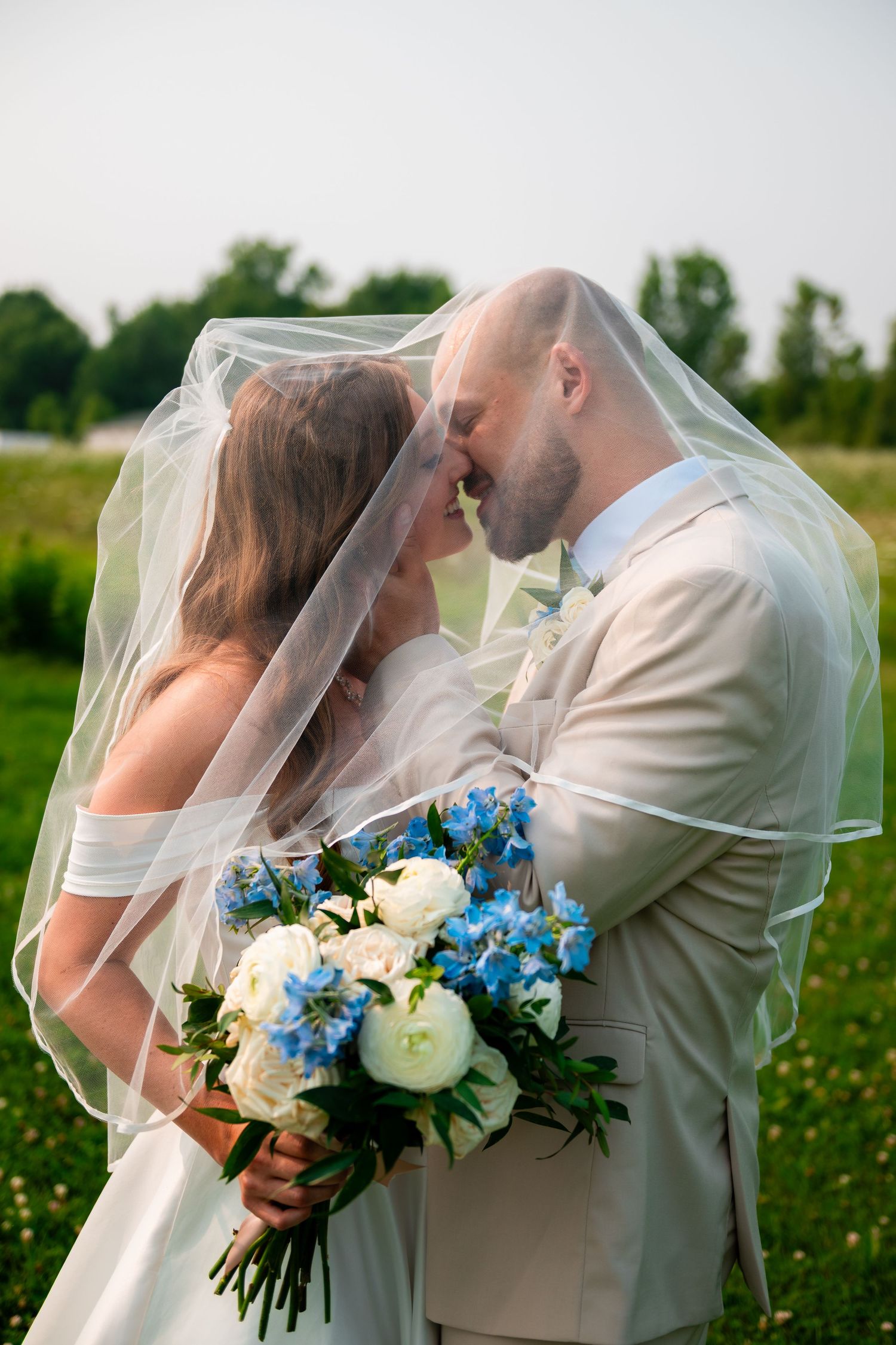 Bride and groom sharing an intimate moment under a veil, holding a white and blue bouquet in a green field.
