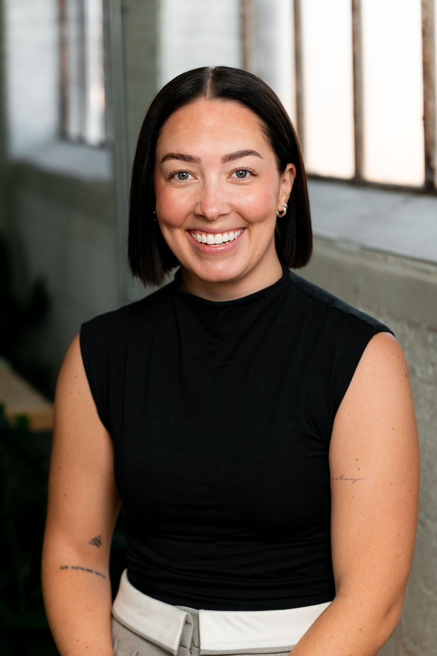 Professional headshot of a person wearing a black sleeveless top against a bright interior background.