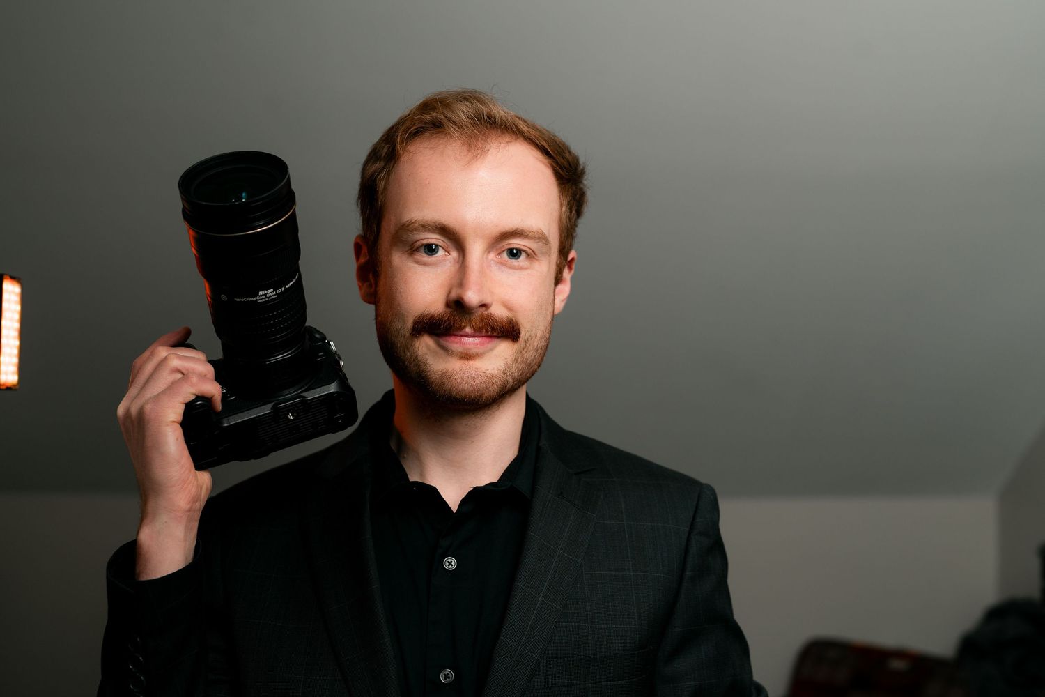 Professional photographer wearing black shirt holds camera equipment against gray studio backdrop.