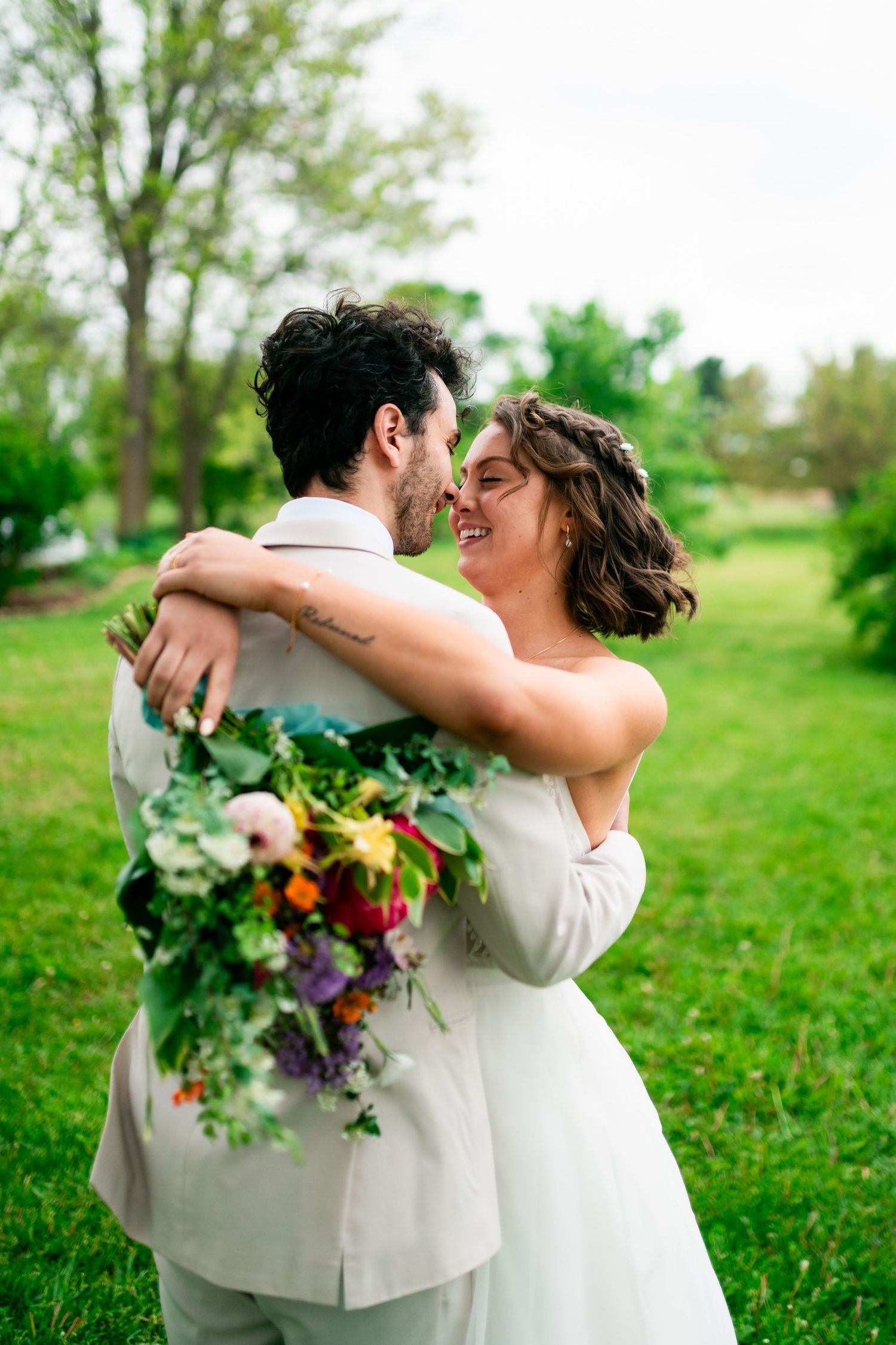 A romantic wedding couple embraces outdoors in a green field with the bride holding a colorful wildflower bouquet.
