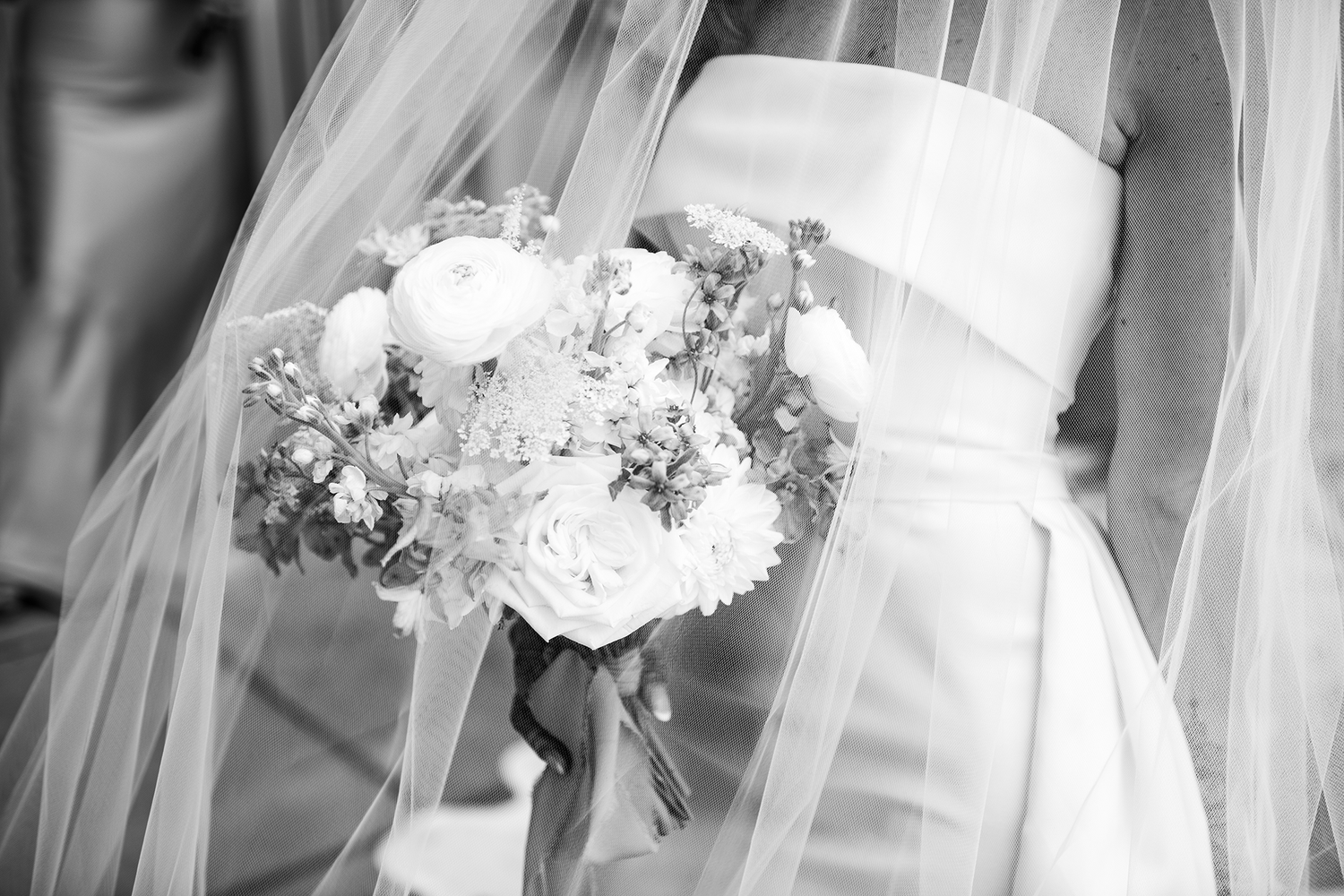 Black and white wedding photo with a sheer veil and white flower bouquet against a white dress.