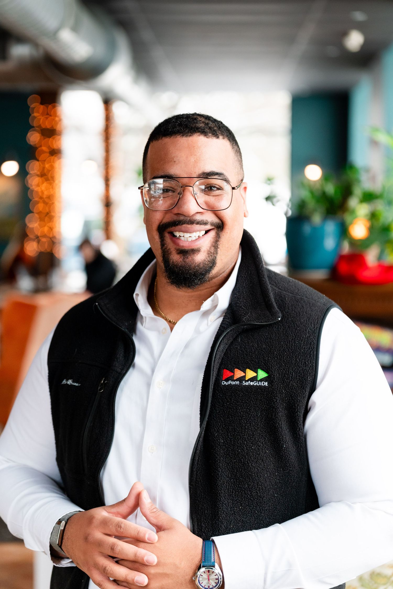 Professional wearing glasses and a black fleece vest with logo smiles warmly in an office setting with decorative lighting.