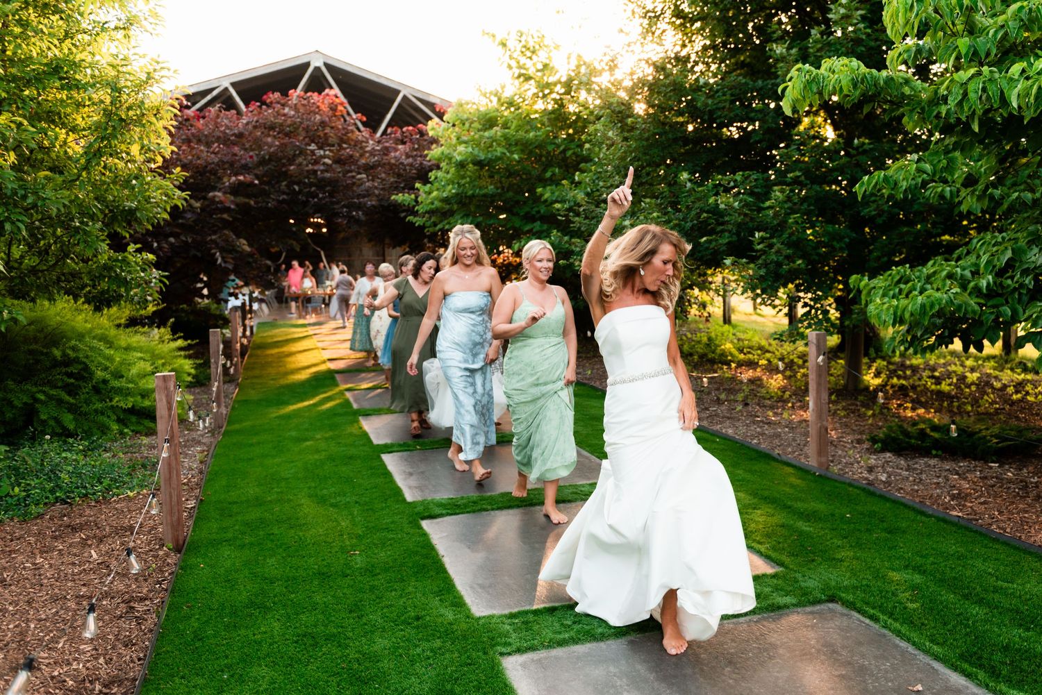 A line of people dancing joyfully down a garden path at an outdoor wedding celebration during golden hour.