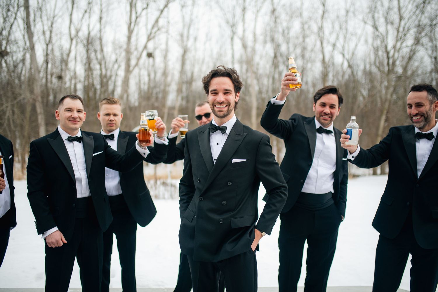 Groom and groomsmen in black tuxedos toasting with drinks outdoors in a snowy winter wedding celebration.