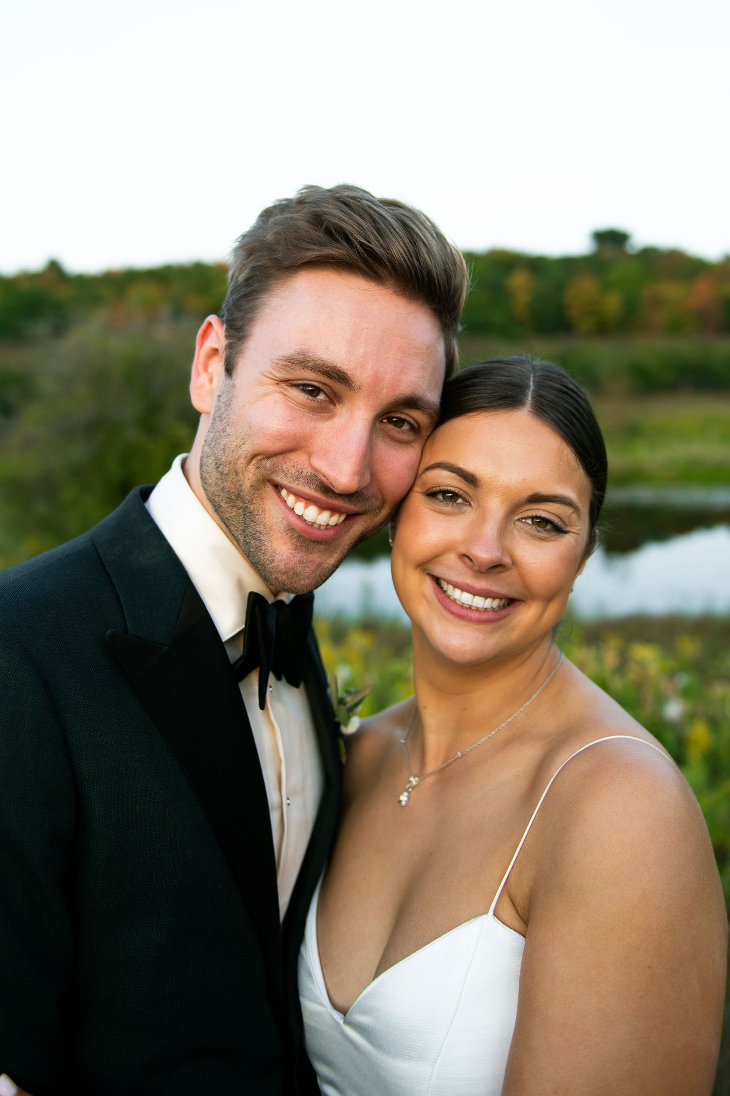 Smiling bride and groom in wedding attire pose cheek to cheek outdoors near a pond with autumn foliage.