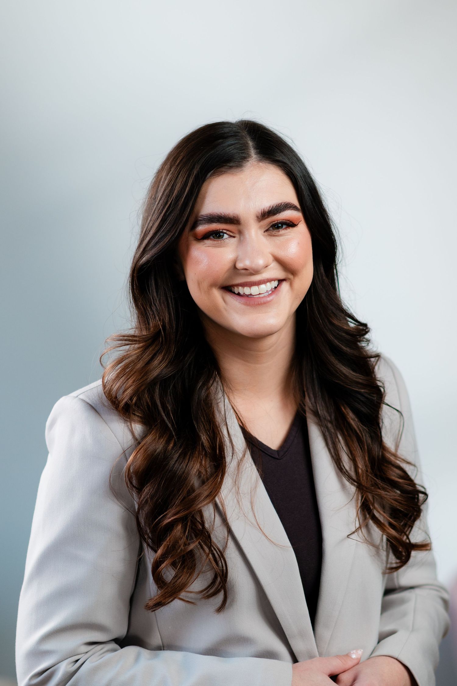Professional headshot portrait of someone wearing a gray blazer and dark top with long wavy brown hair against gray backdrop in Michigan