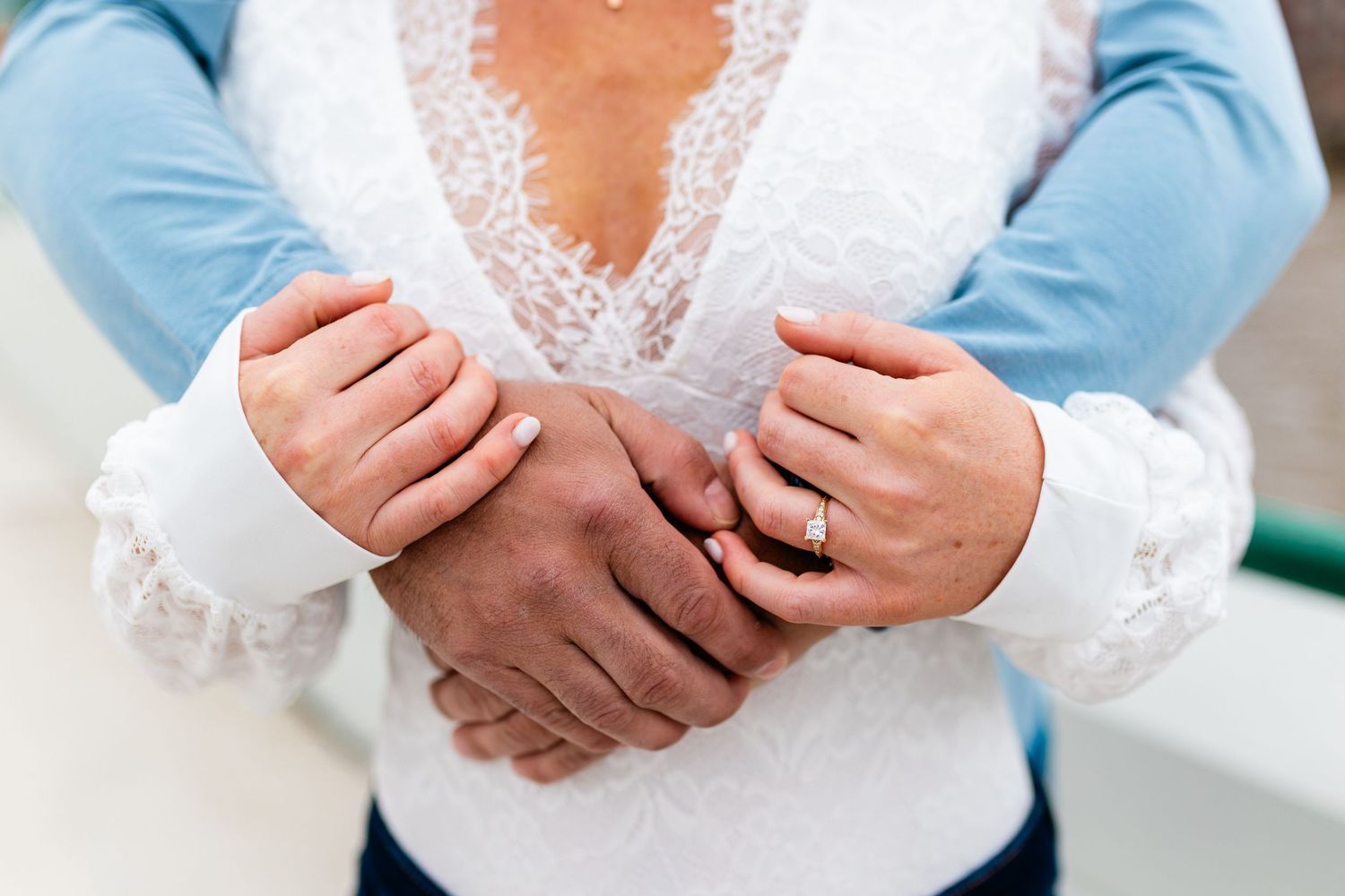 Close-up of couple holding hands showing engagement ring, bride in white lace dress embraced from behind.